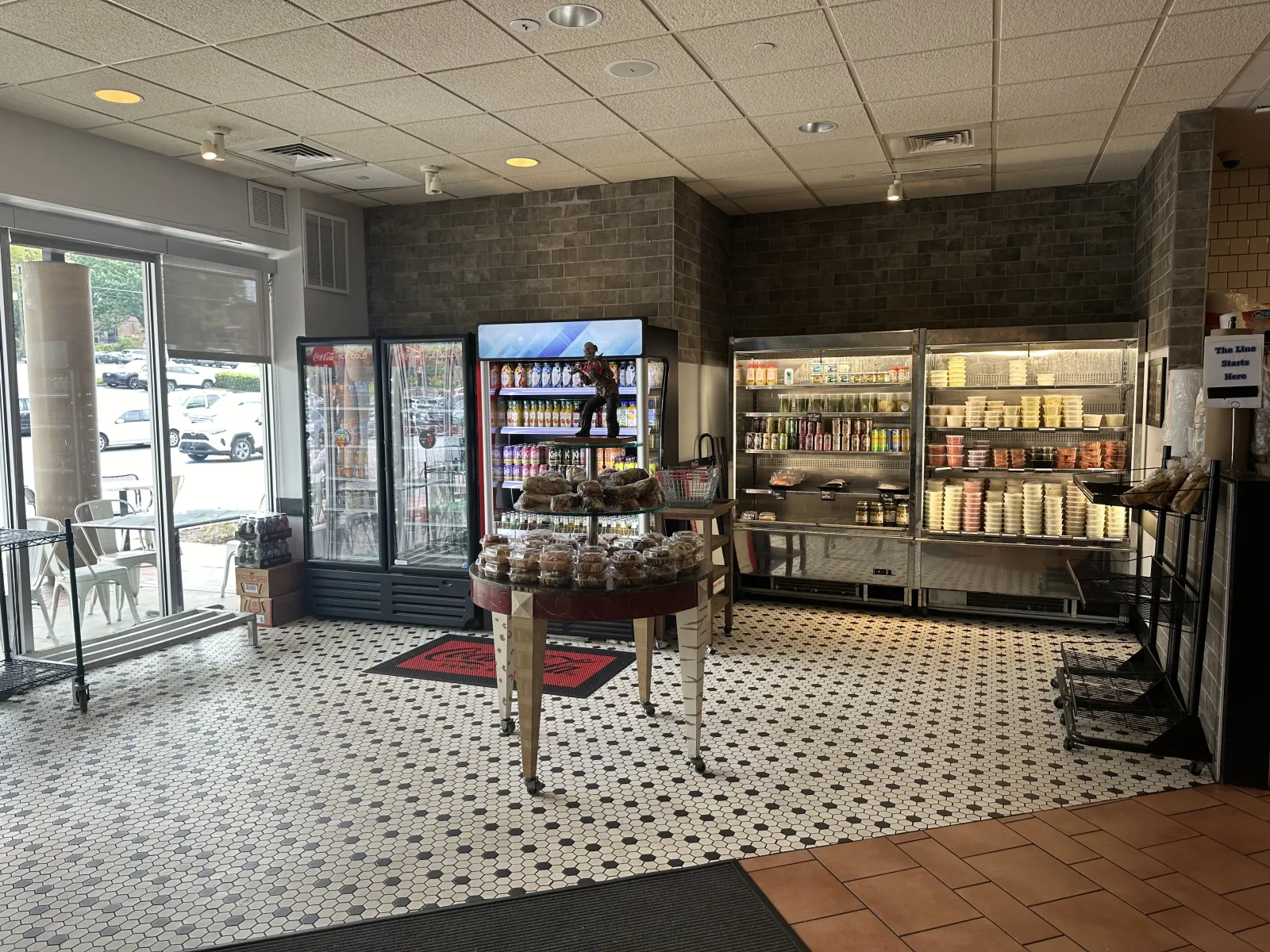 Interior of a deli with refrigerated display cases, shelves of food products, and a central table with baked goods.