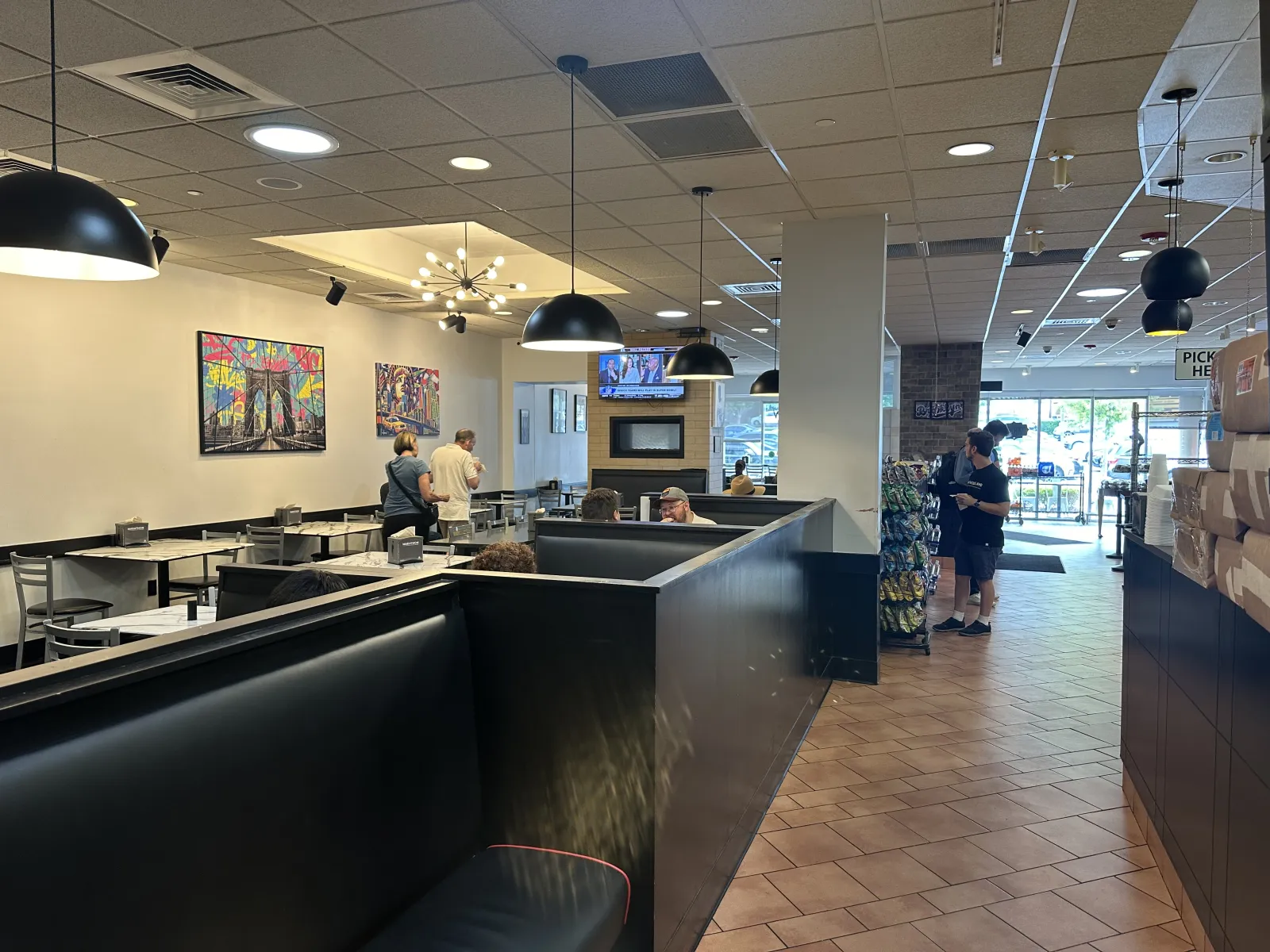 Interior of a casual restaurant with black booth seating, hanging lamps, colorful wall art, and tile floors.