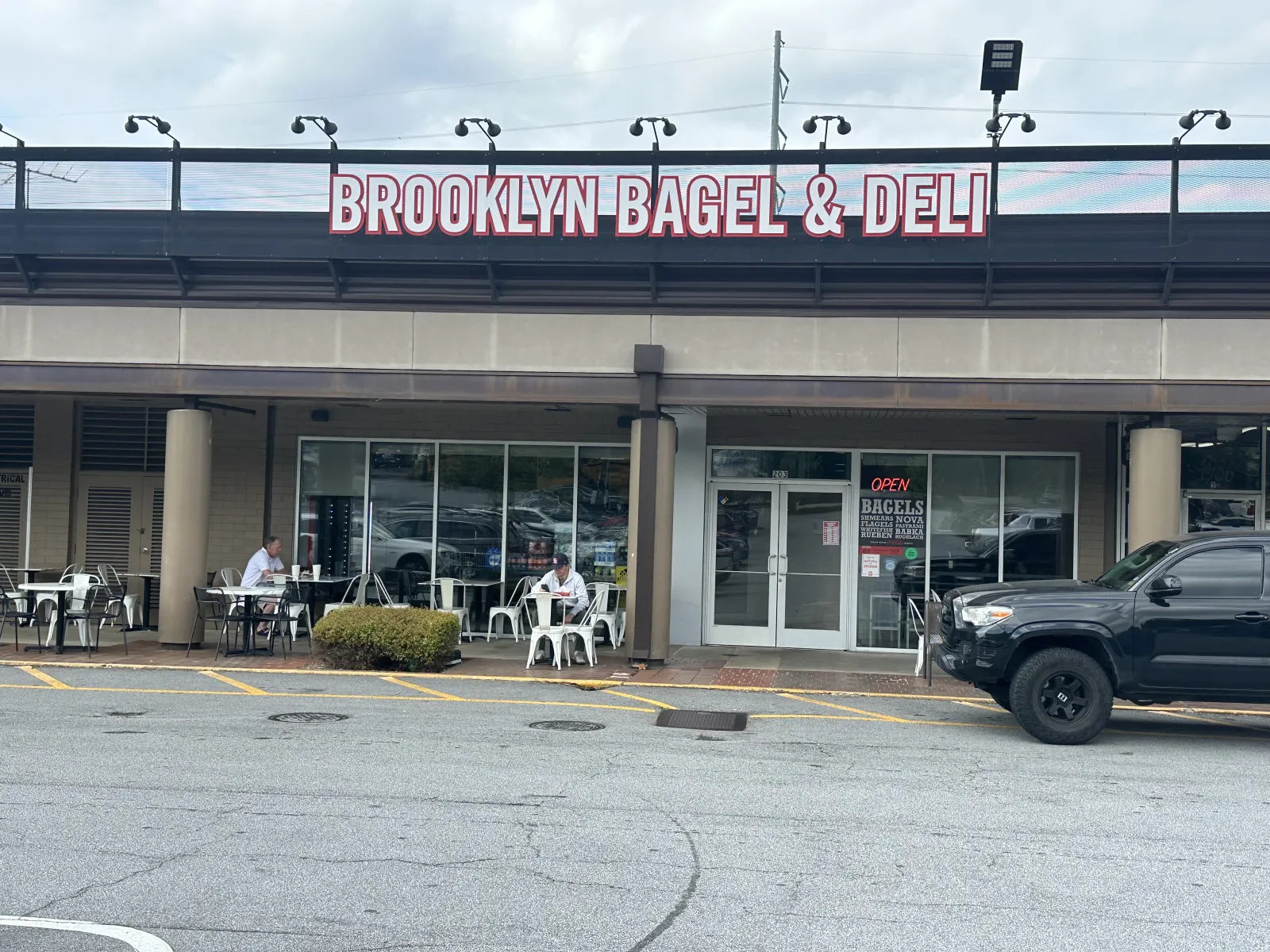 Brooklyn Bagel & Deli storefront with outdoor seating and customers, black truck parked nearby.