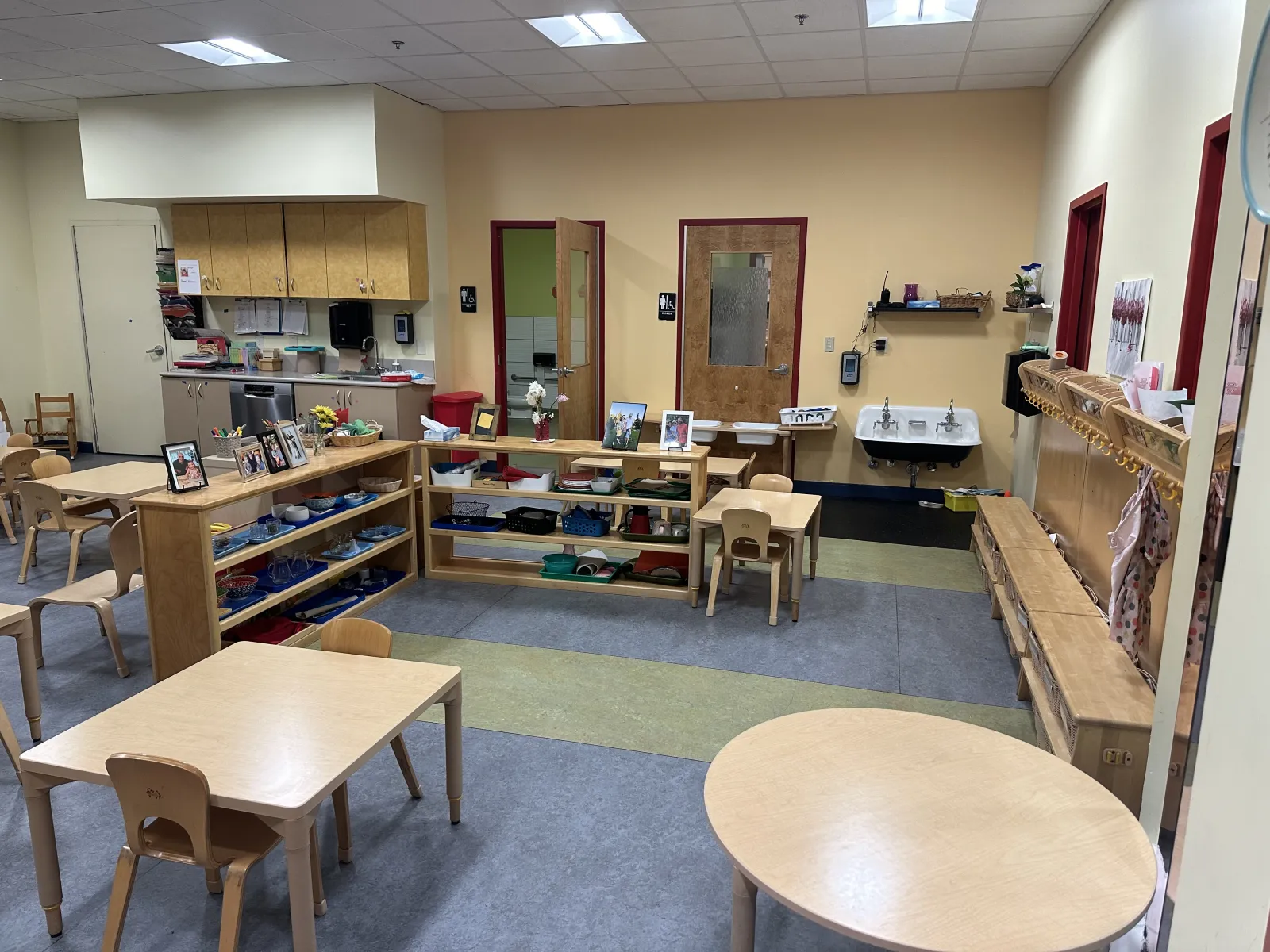 Empty preschool classroom with small wooden tables, chairs, shelves, and art supplies neatly organized.