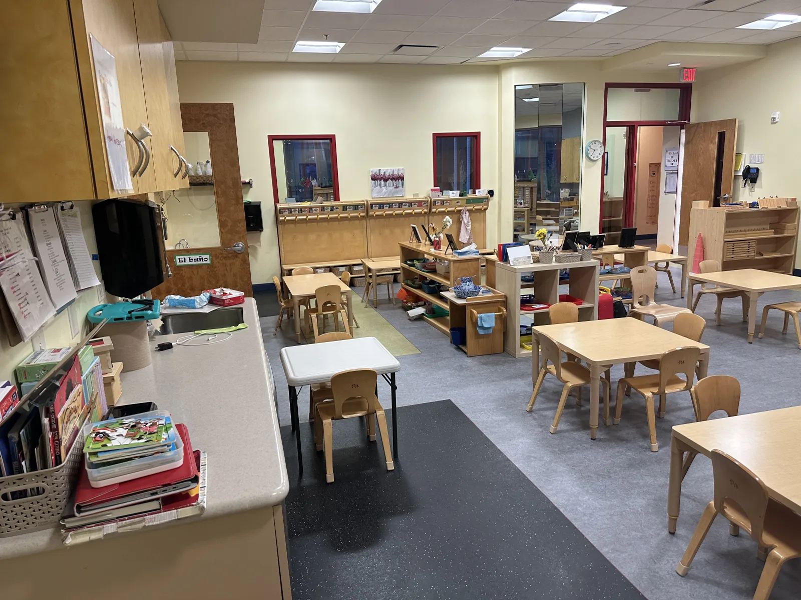 Bright preschool classroom with small wooden tables, chairs, shelves, and educational materials.