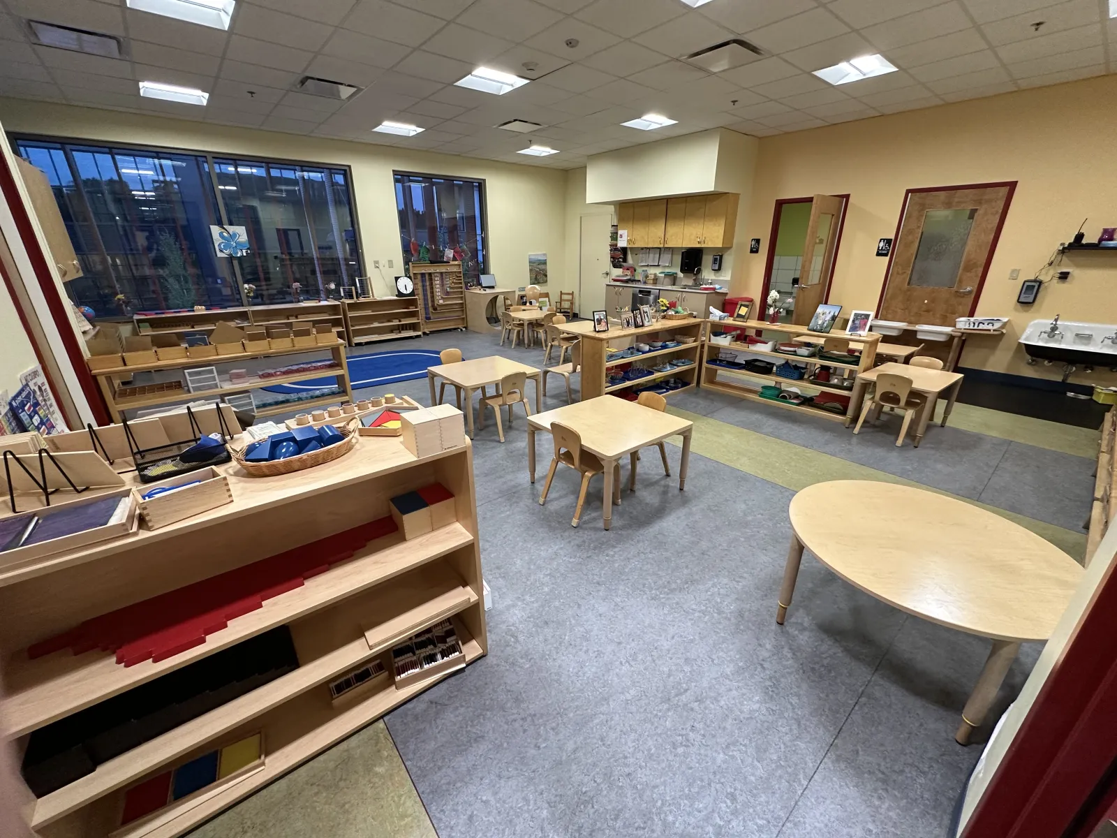 Spacious Montessori classroom with wooden tables, chairs, learning materials, and large windows for natural light.