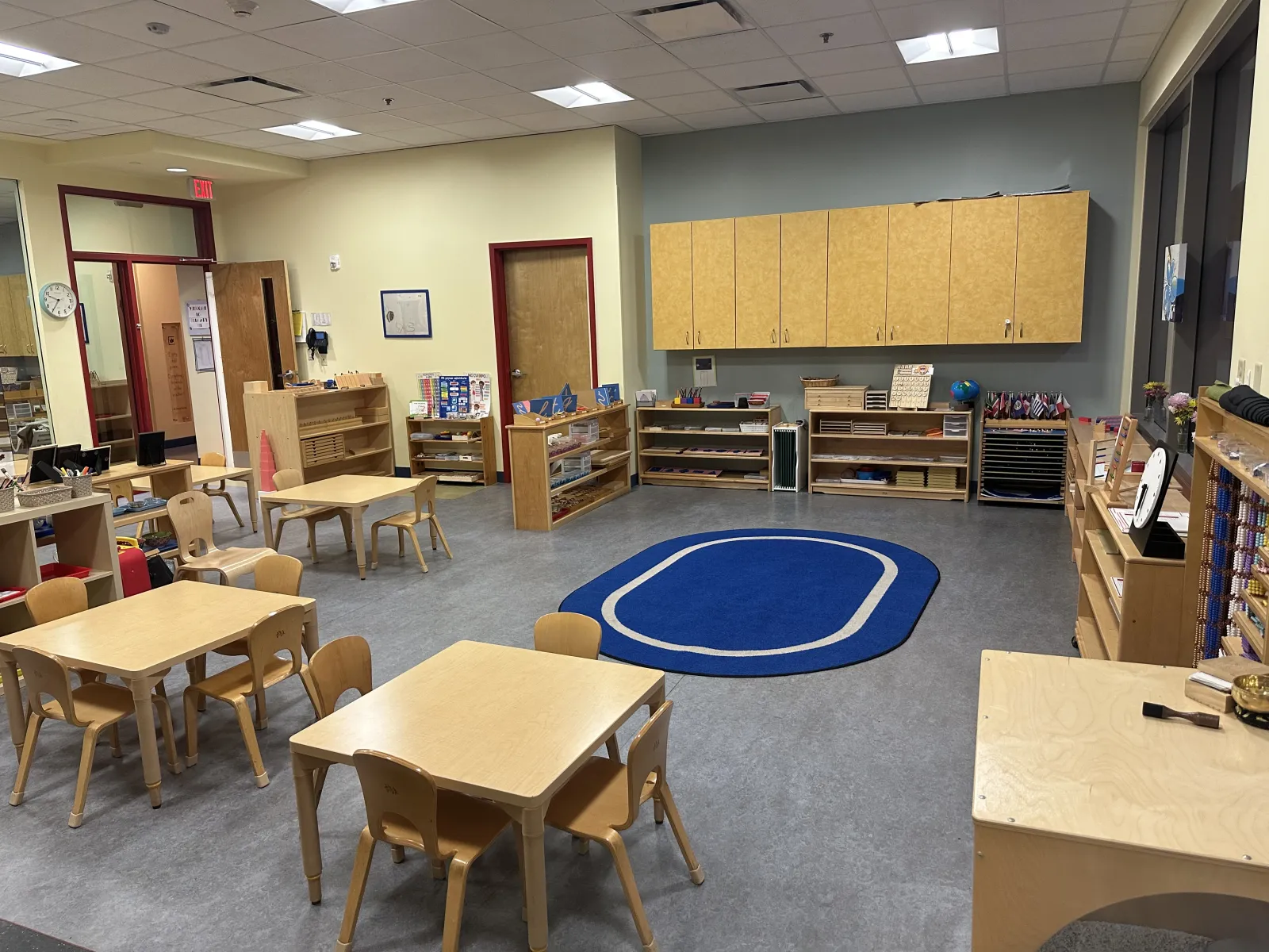 Bright and organized classroom with wooden tables, chairs, shelves, and a blue oval rug on gray floor.