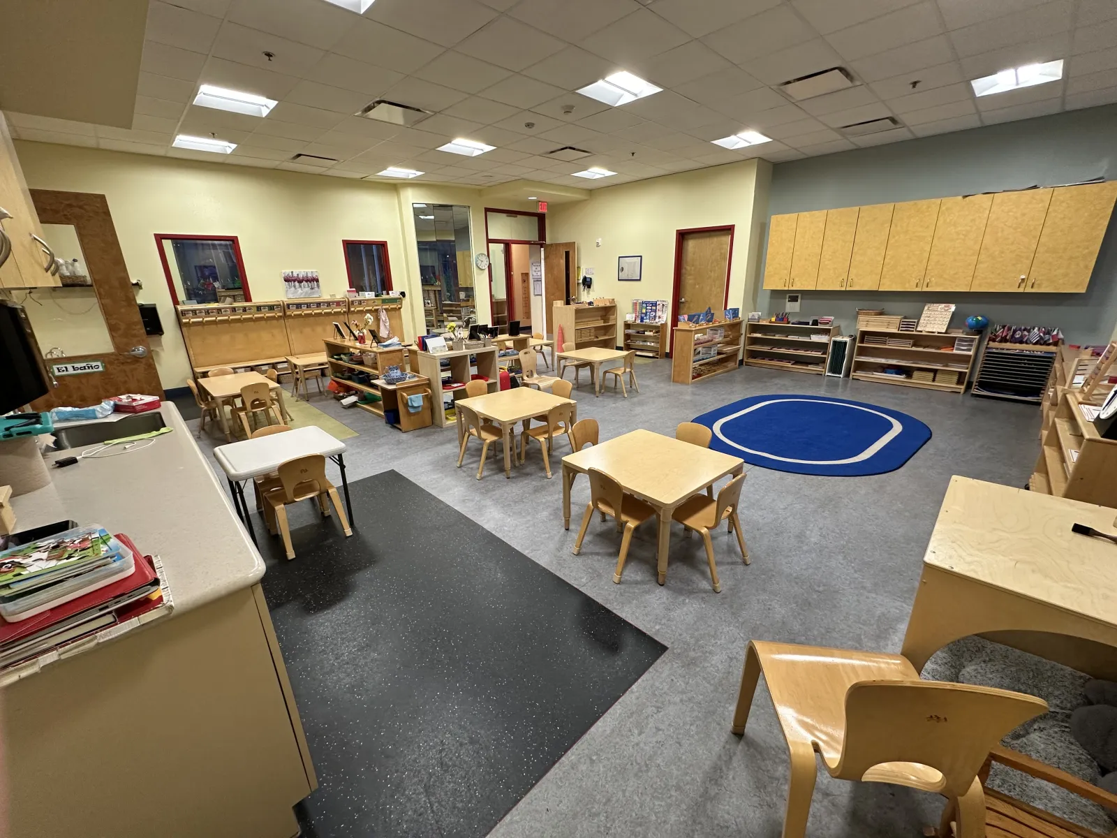 Spacious preschool classroom with small tables, chairs, shelves, and a blue carpet area for group activities.