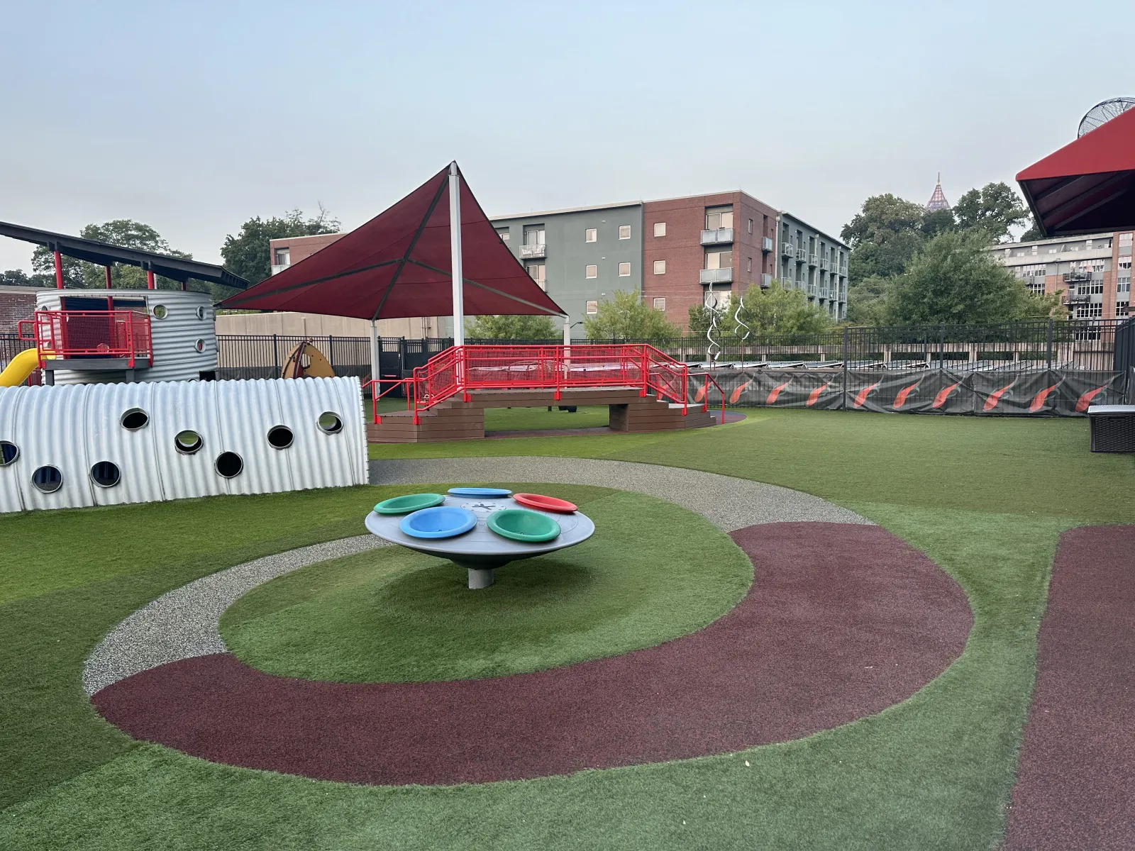 Modern playground with colorful round play panels, shade canopy, and soft turf in an urban setting.
