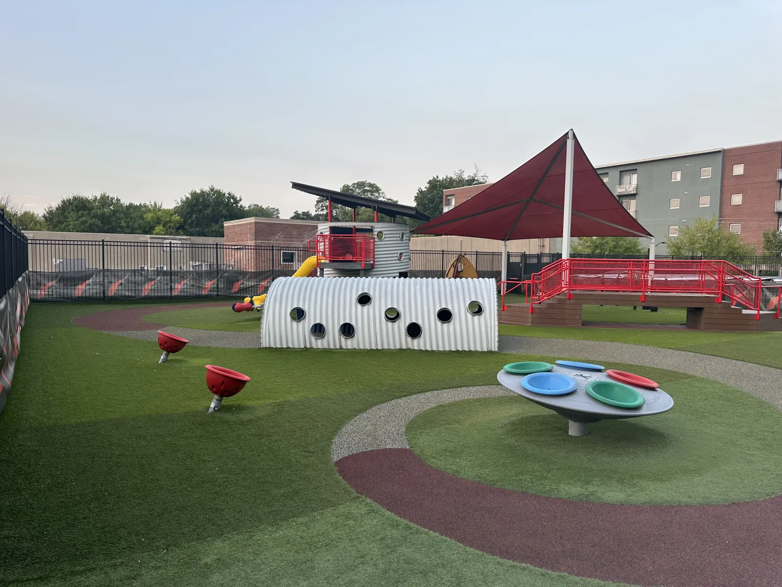 Modern outdoor playground with colorful spinning discs, slide, climbing structures, and shaded red canopy over seating area.