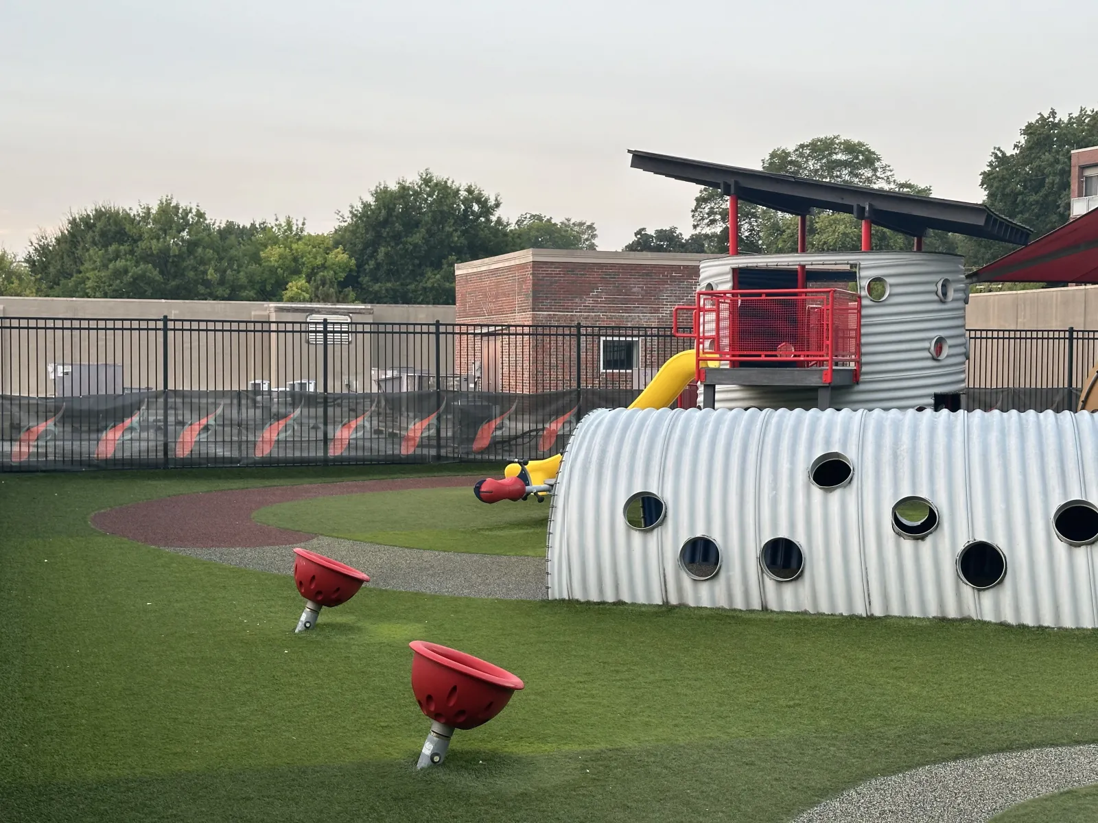 Modern playground with metal tunnel, red spinning seats, yellow slide, and turf surface under overcast sky