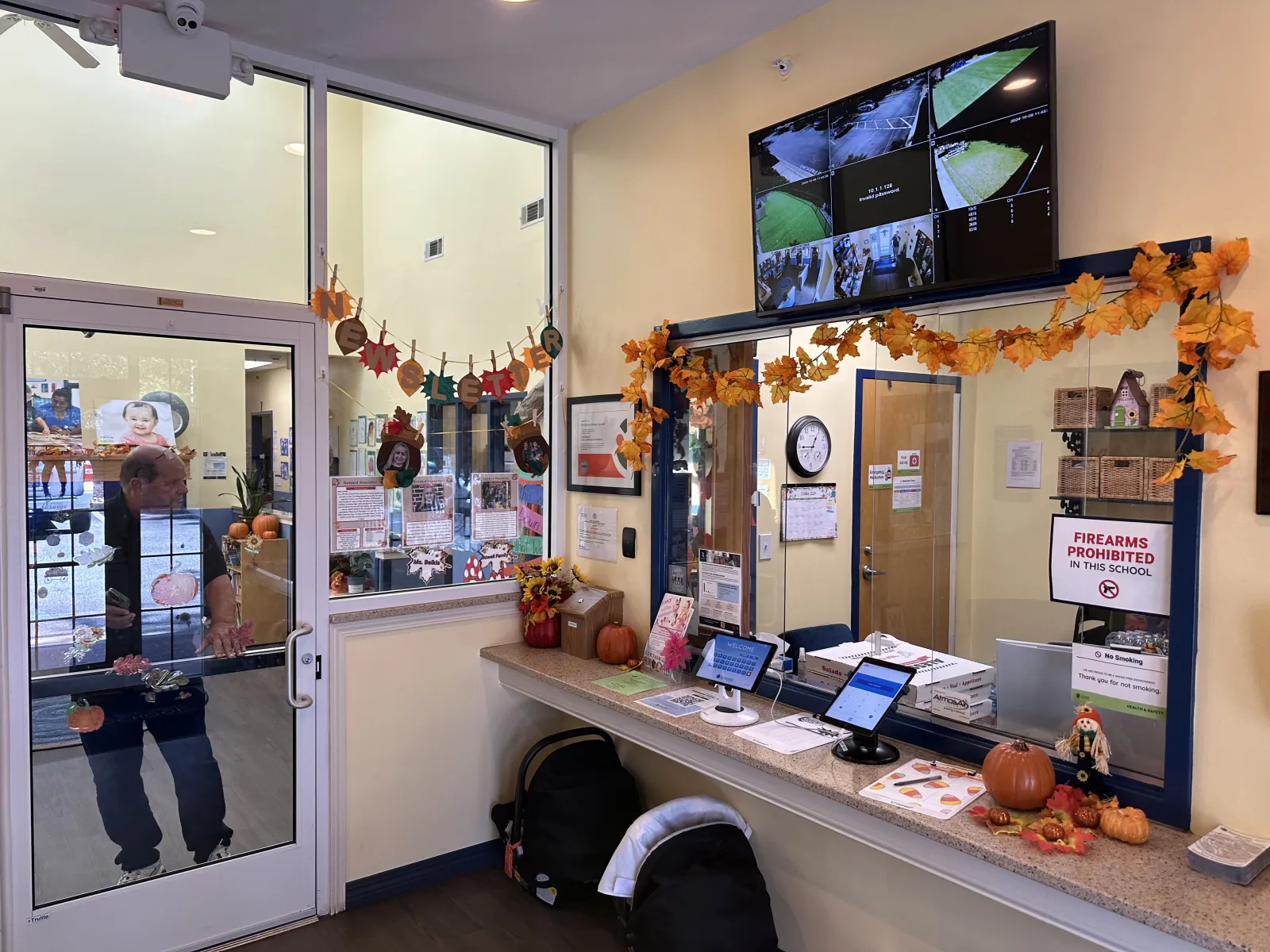 School office entrance decorated with autumn leaves, pumpkins, security monitors, and visitor check-in tablets.