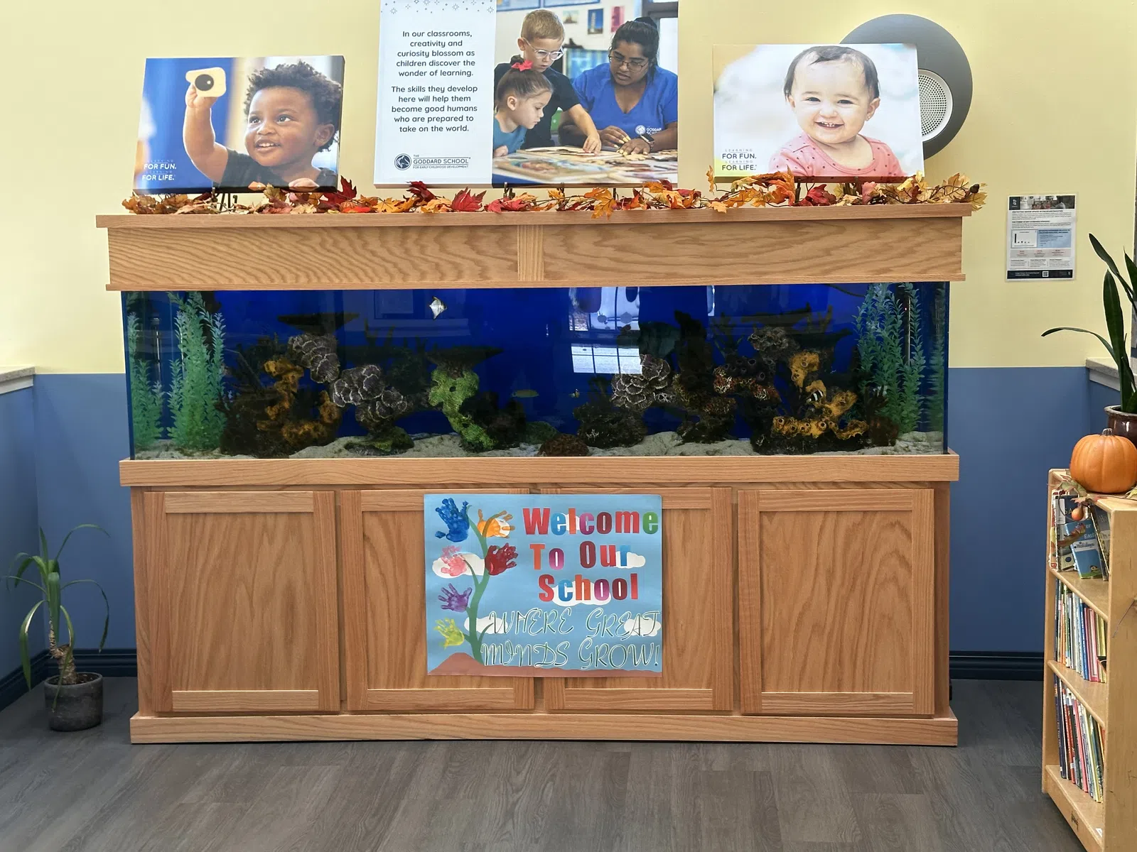 Large aquarium with decorative plants in a school lobby featuring welcome signs and children's photos above it.