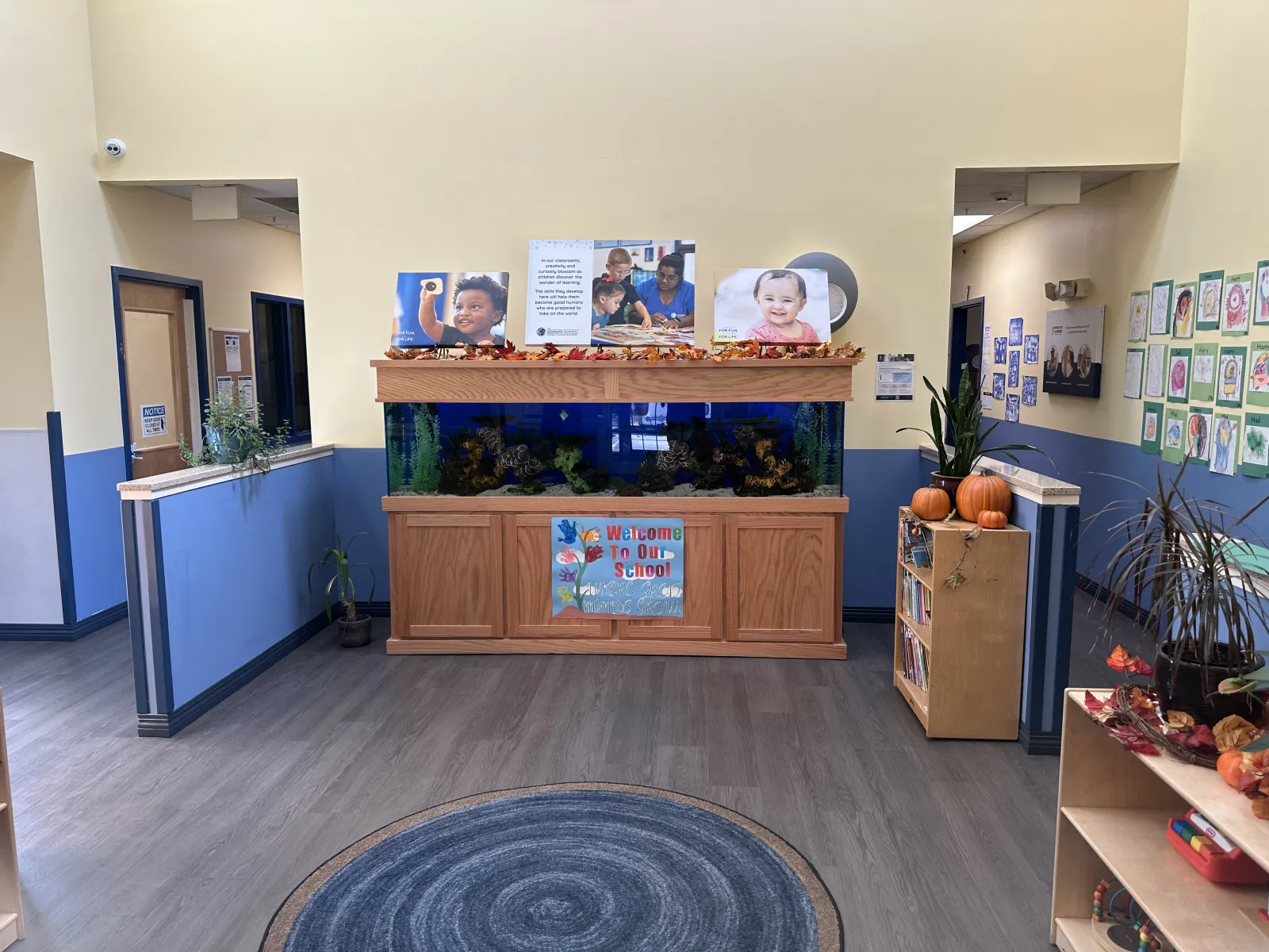 School lobby with large aquarium, welcome sign, children's artwork, bookshelves, and autumn decorations including pumpkins.