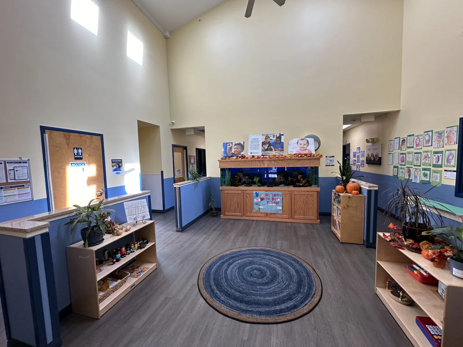 Bright, spacious daycare lobby with blue circular rug, fish tank, shelves with toys, and children's artwork on walls.