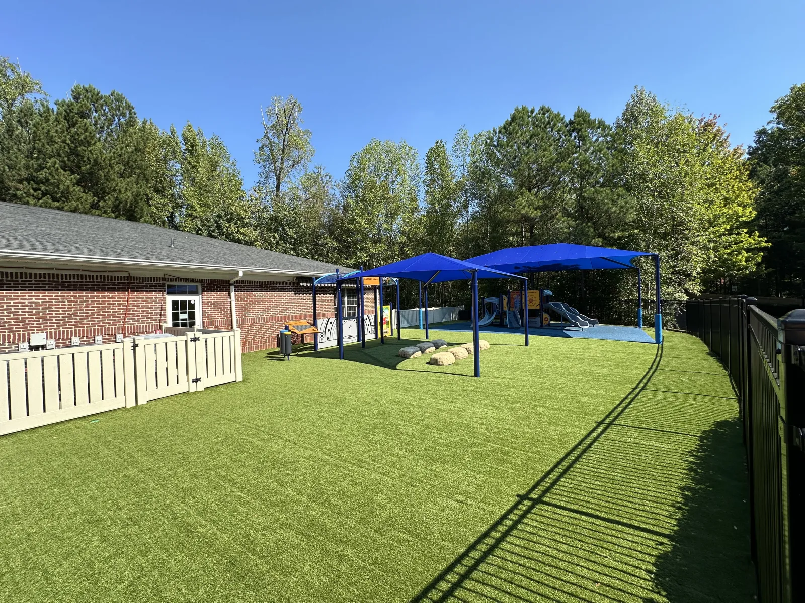 Outdoor playground with artificial turf, blue shade canopies, slides, and a brick building surrounded by trees.