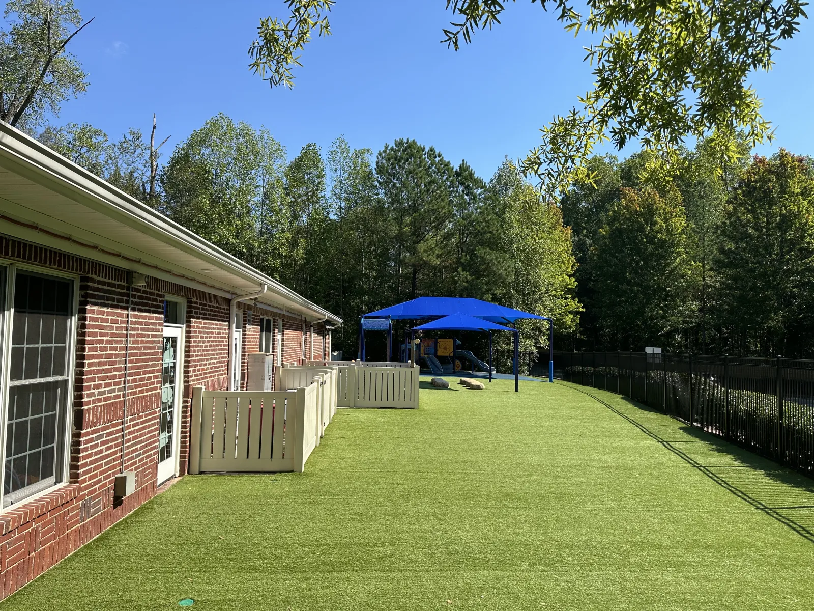 Outdoor playground with artificial turf, blue shade structure, and brick building under clear blue sky.