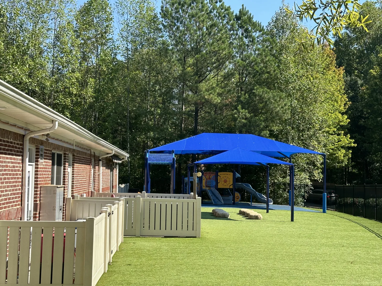 Outdoor playground with blue shade sails, artificial grass, play structures, and fenced area next to brick building.