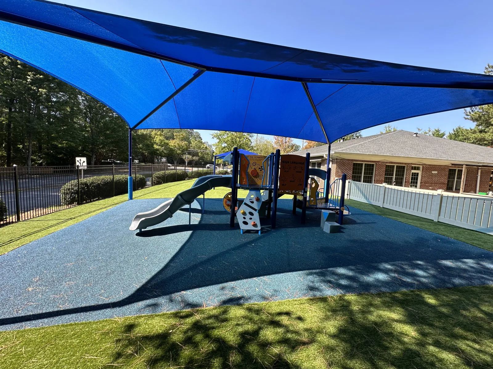 Outdoor playground with slides and climbing structures shaded by large blue canopy on sunny day.
