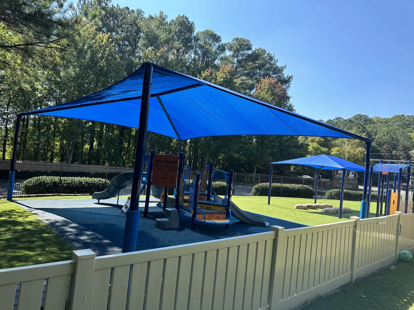 Children's playground with blue shade structures, slides, and climbing equipment surrounded by trees and white fence.