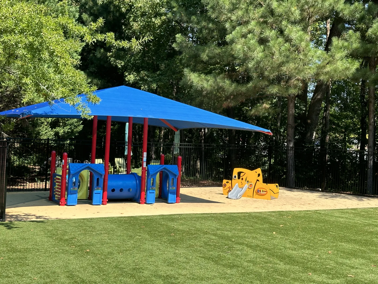 Children's playground with blue shade canopy, colorful play structures, slides, and artificial grass surrounded by trees.