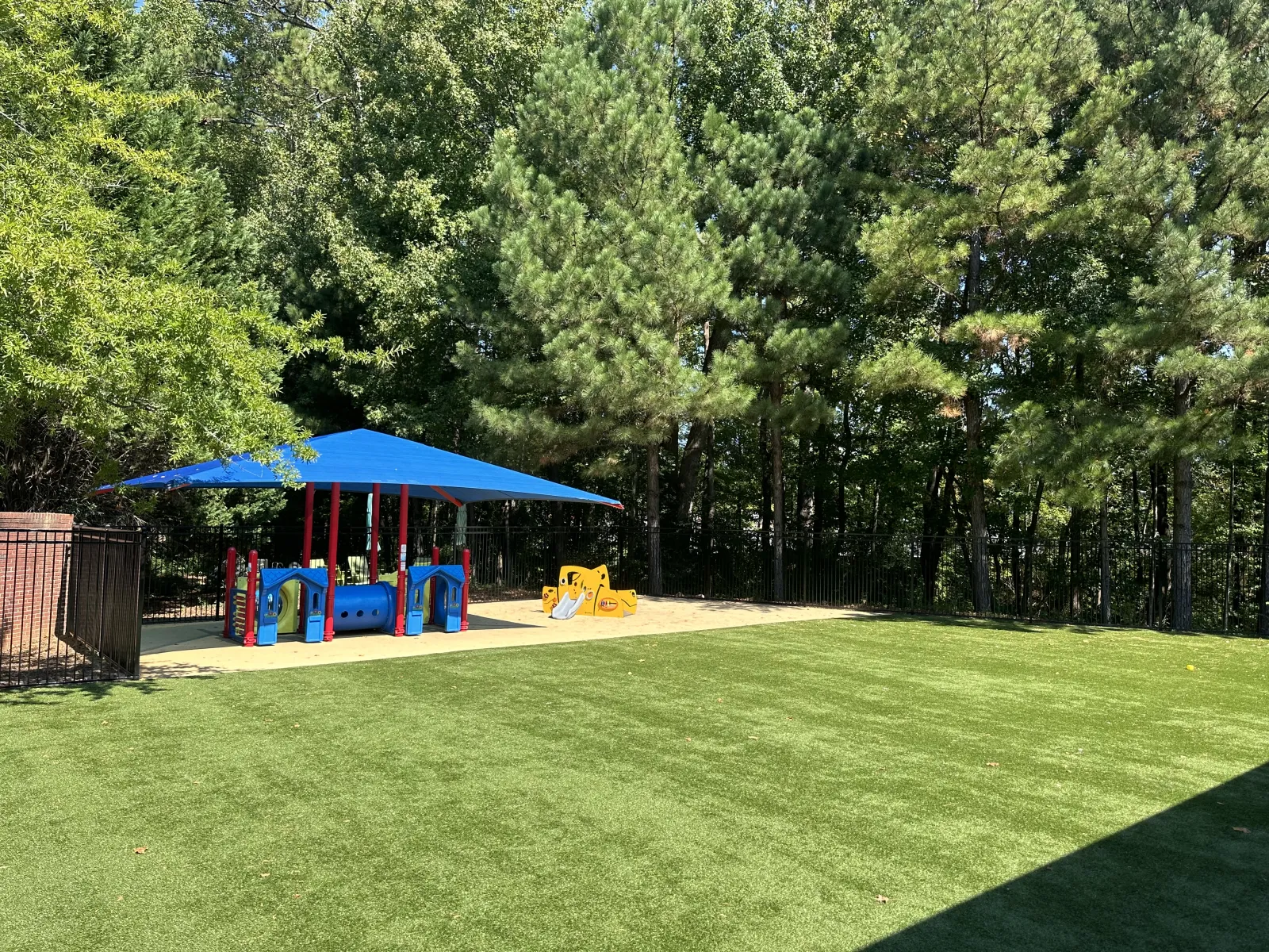 Children's playground with blue shade cover, colorful play structures, and green grassy area surrounded by trees.