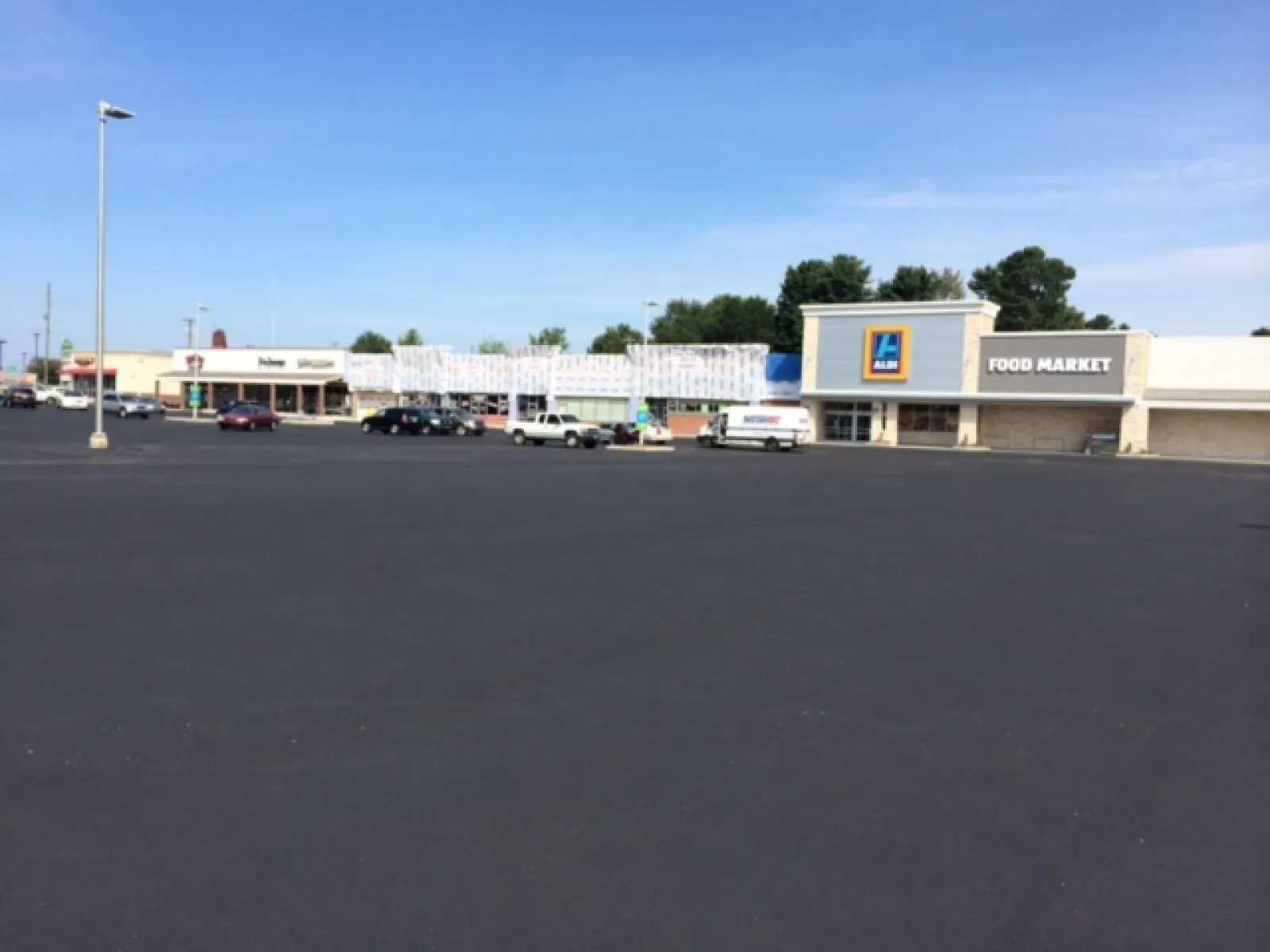 Wide empty parking lot in front of a strip mall featuring Aldi Food Market and other stores under clear blue sky