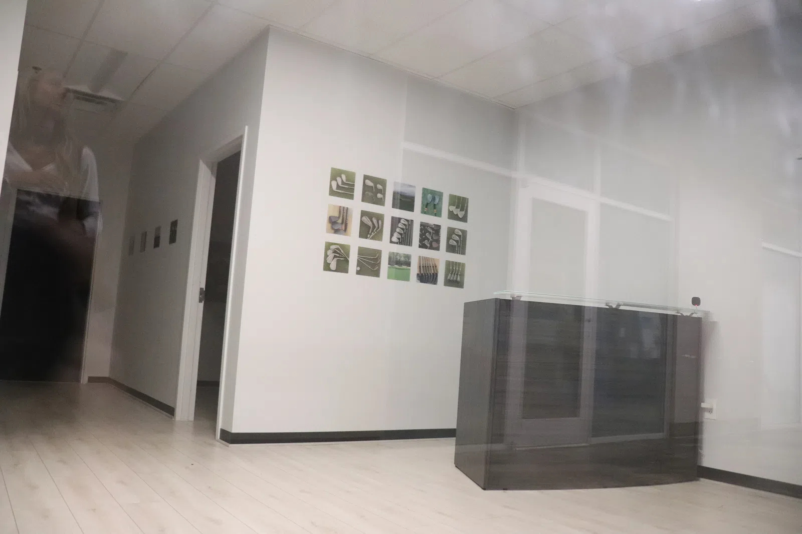 Modern office hallway with light wood flooring, white walls, and a dark reception desk under soft ceiling lights