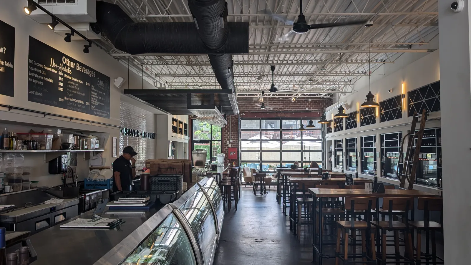Interior of a modern cafe with high ceilings, wooden tables, industrial decor, and a glass display counter.
