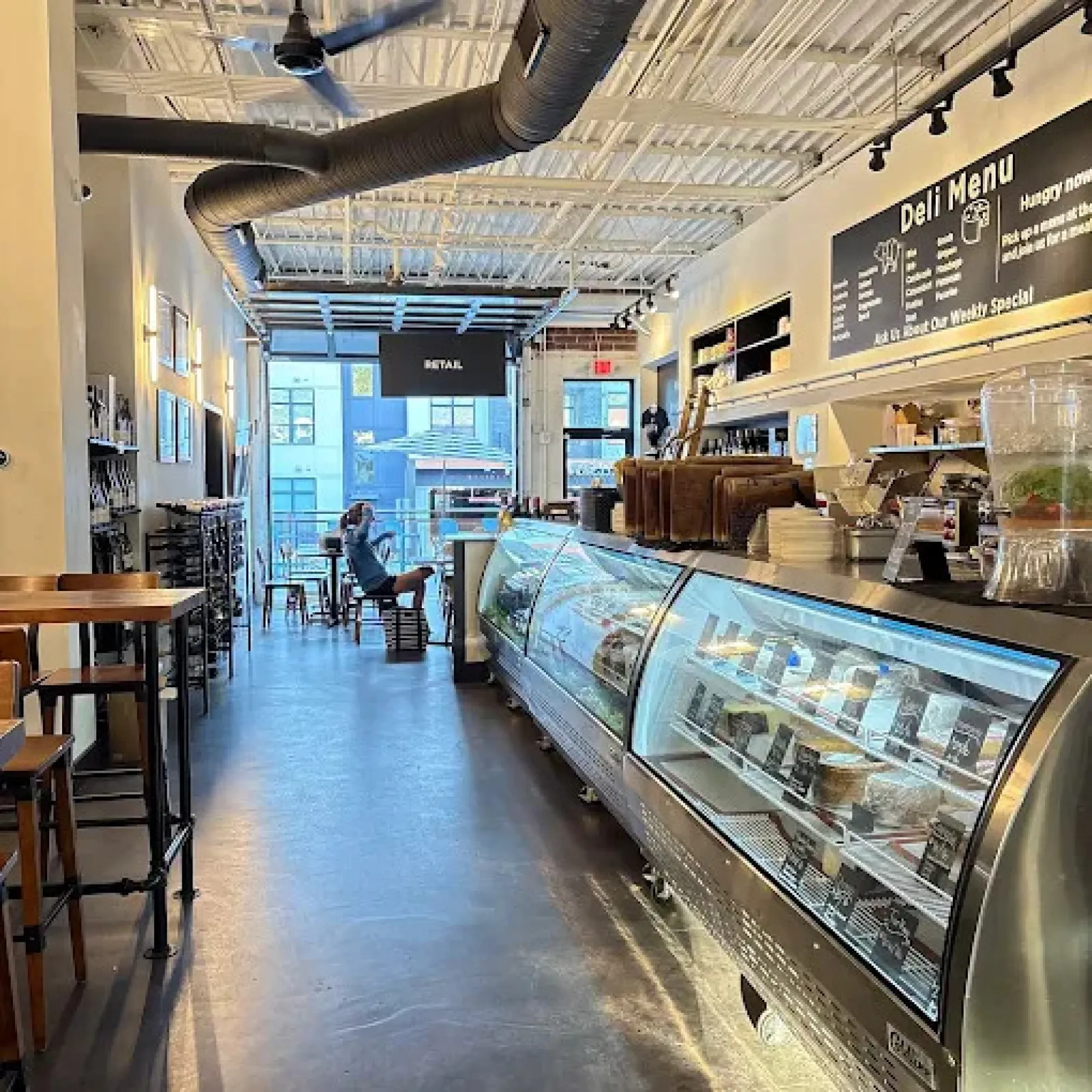 Bright modern deli interior with glass display cases, seating area, and a person sitting near the entrance.