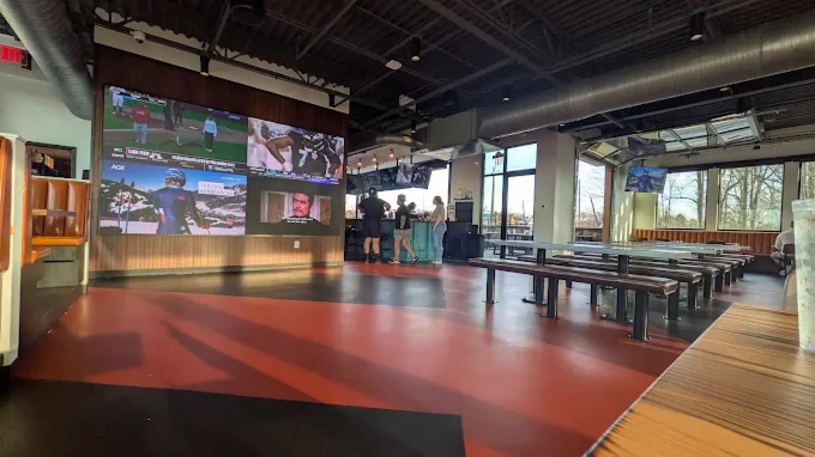Modern sports bar with large screens showing games, industrial ceiling, wooden benches, and people near the bar counter.