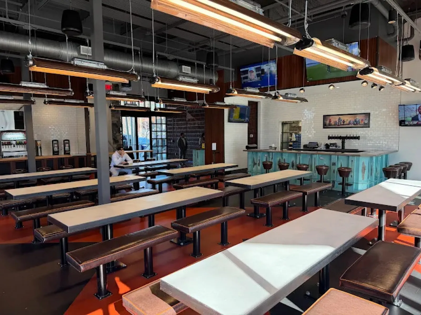 Modern industrial-style cafeteria with long tables, bench seating, hanging lights, and a bar area in the background