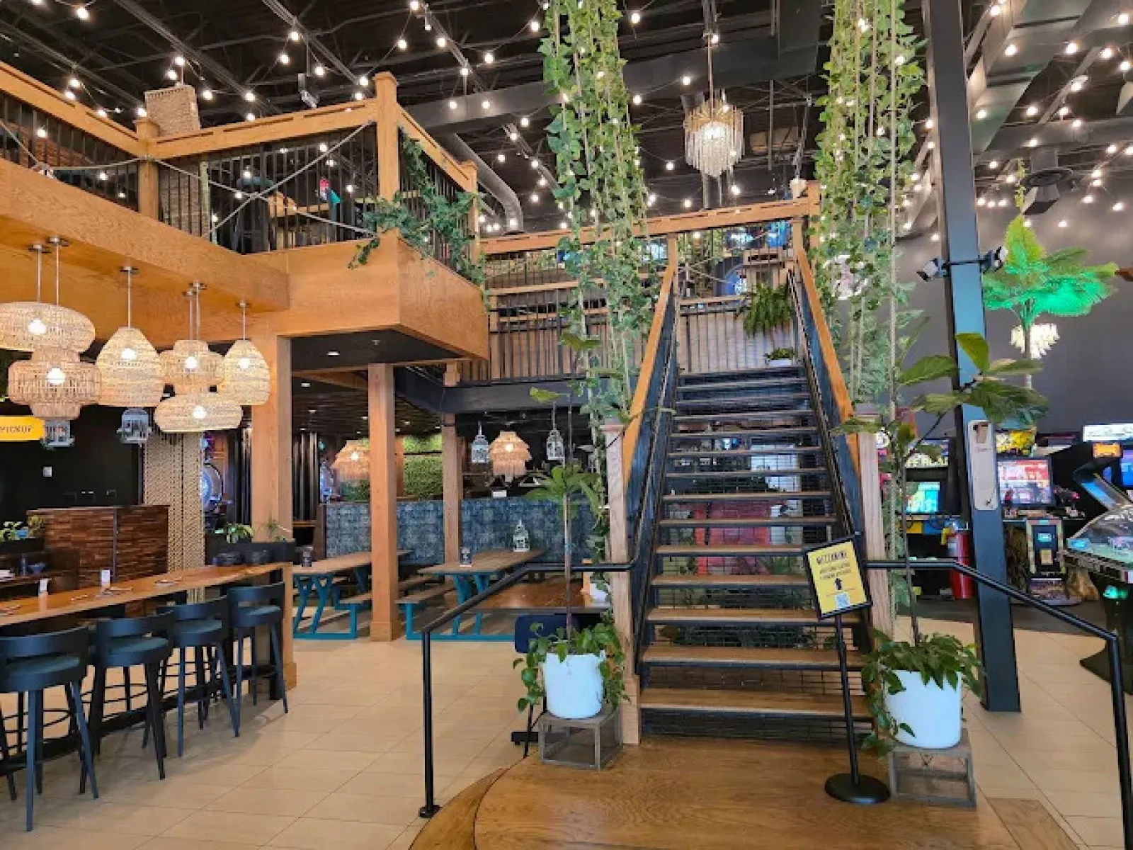 Modern indoor restaurant with wooden tables, hanging lights, plants, staircase, and arcade games in the background