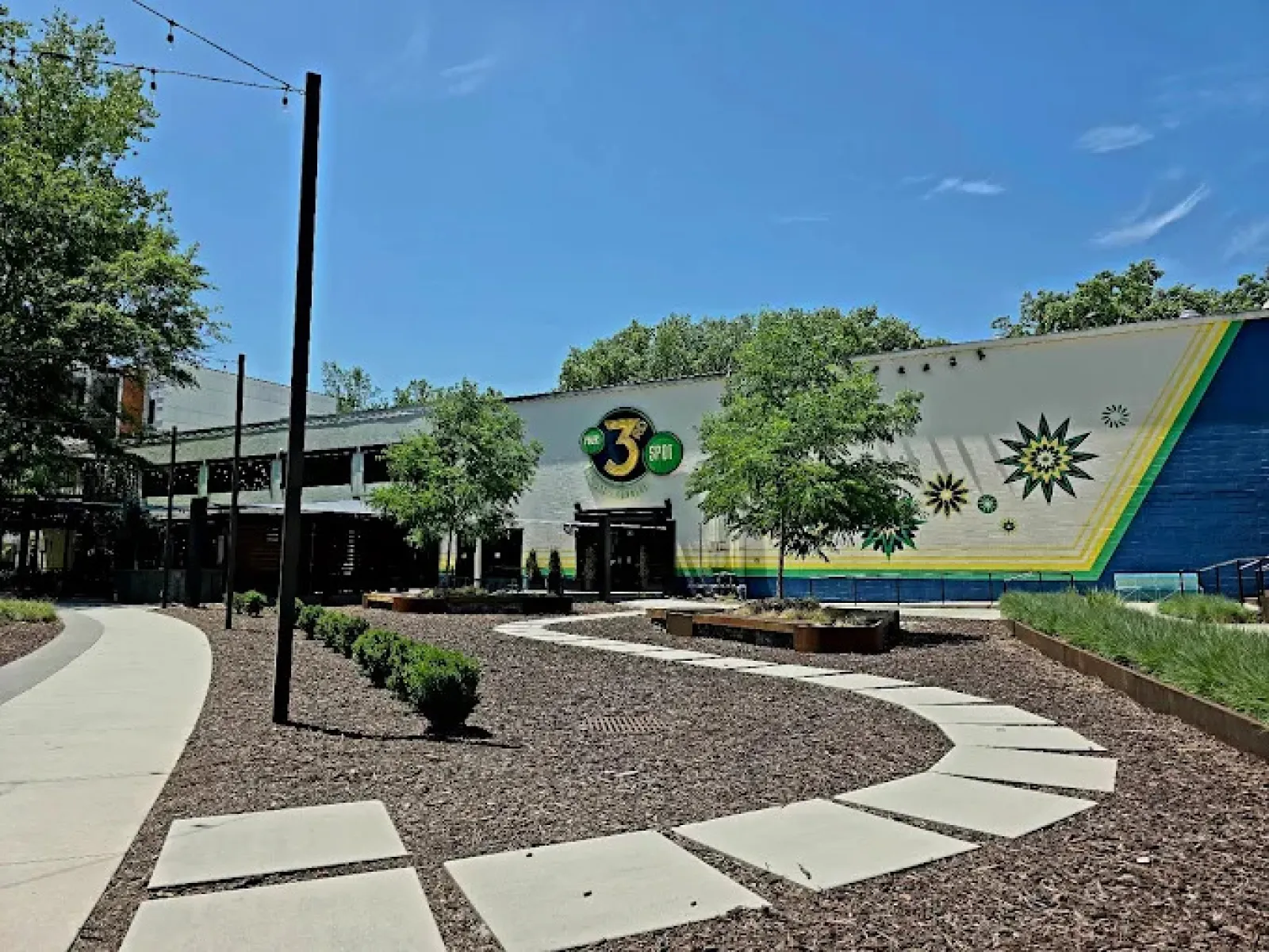Outdoor view of The 3 Spot building with curved sidewalk, trees, and decorative mural under blue sky.
