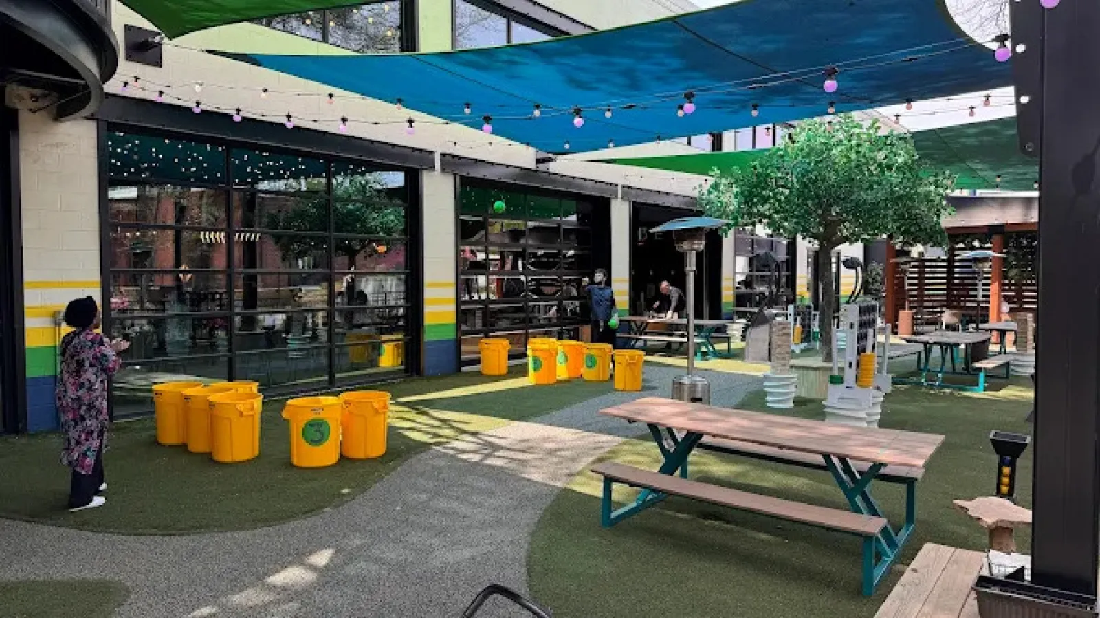 Outdoor seating area with picnic tables, yellow recycling bins, shade sails, and string lights in a commercial space.