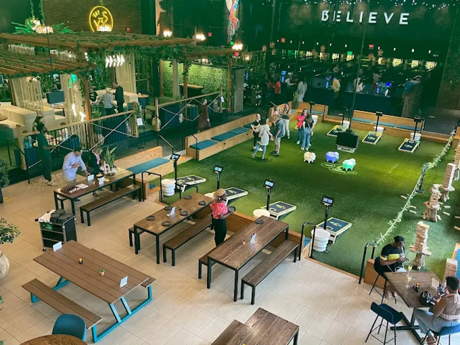 Indoor recreational space with people playing cornhole, sitting at wooden tables, and green turf floors under plants.