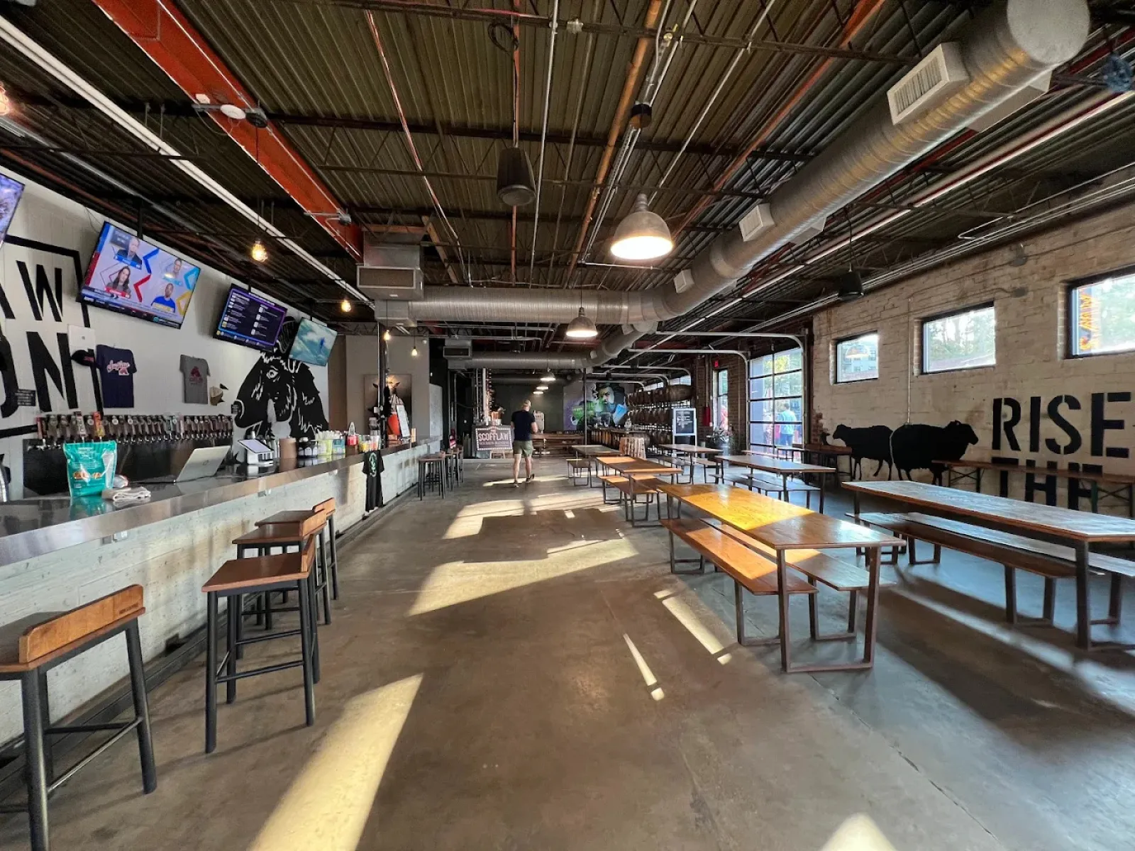 Spacious brewery tasting room with bar stools, wooden tables, industrial ceiling, and wall art of cows and text.