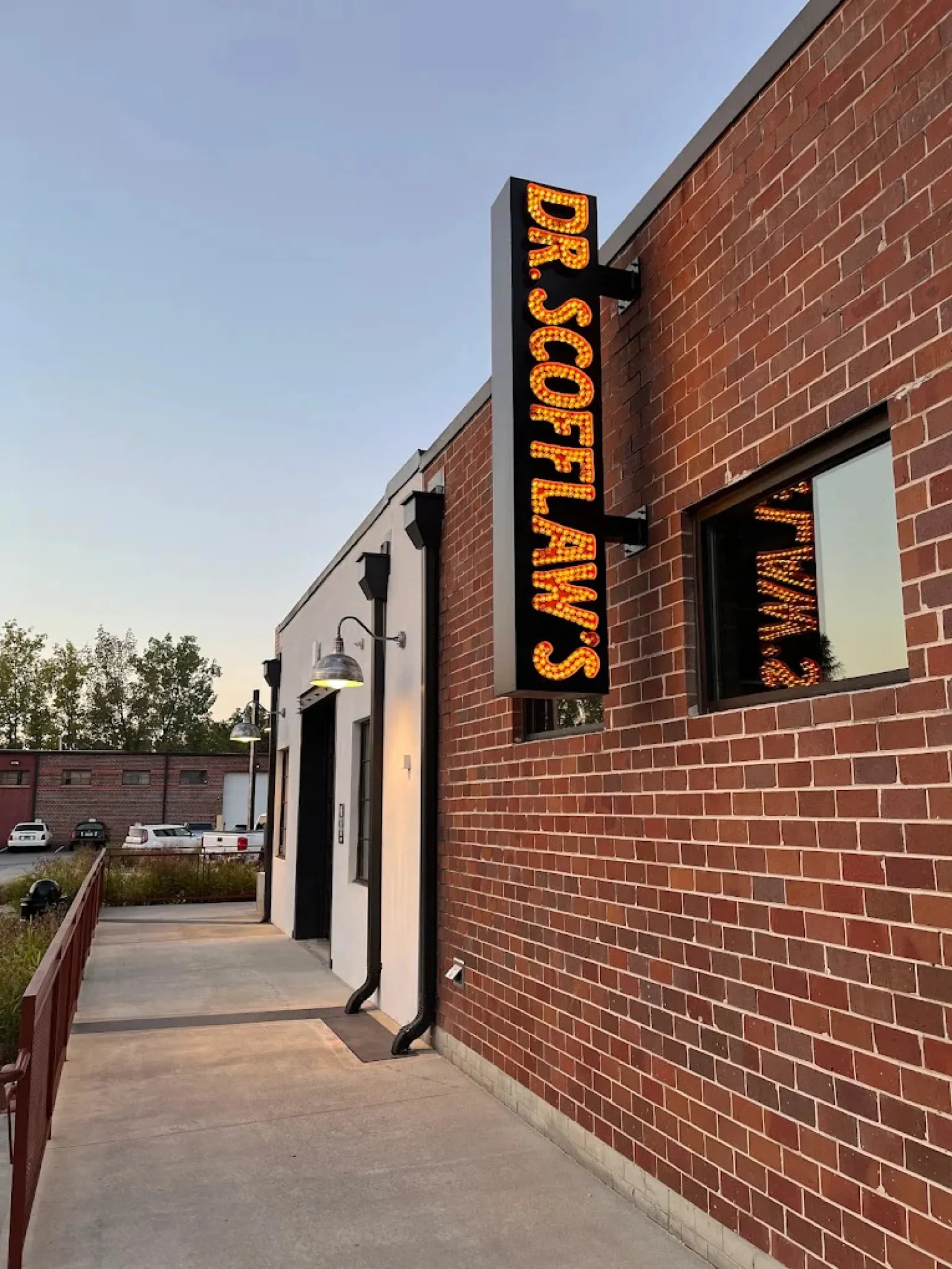 Lit vertical sign reading Dr. Scoflaws mounted on a brick building facade with sidewalk and street lamps.