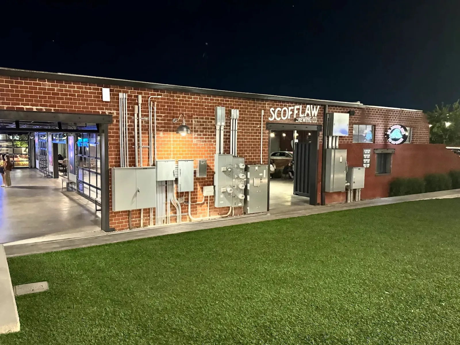 Night view of Scofflaw Brewing Co building with exposed brick, electrical panels, and green lawn in front.