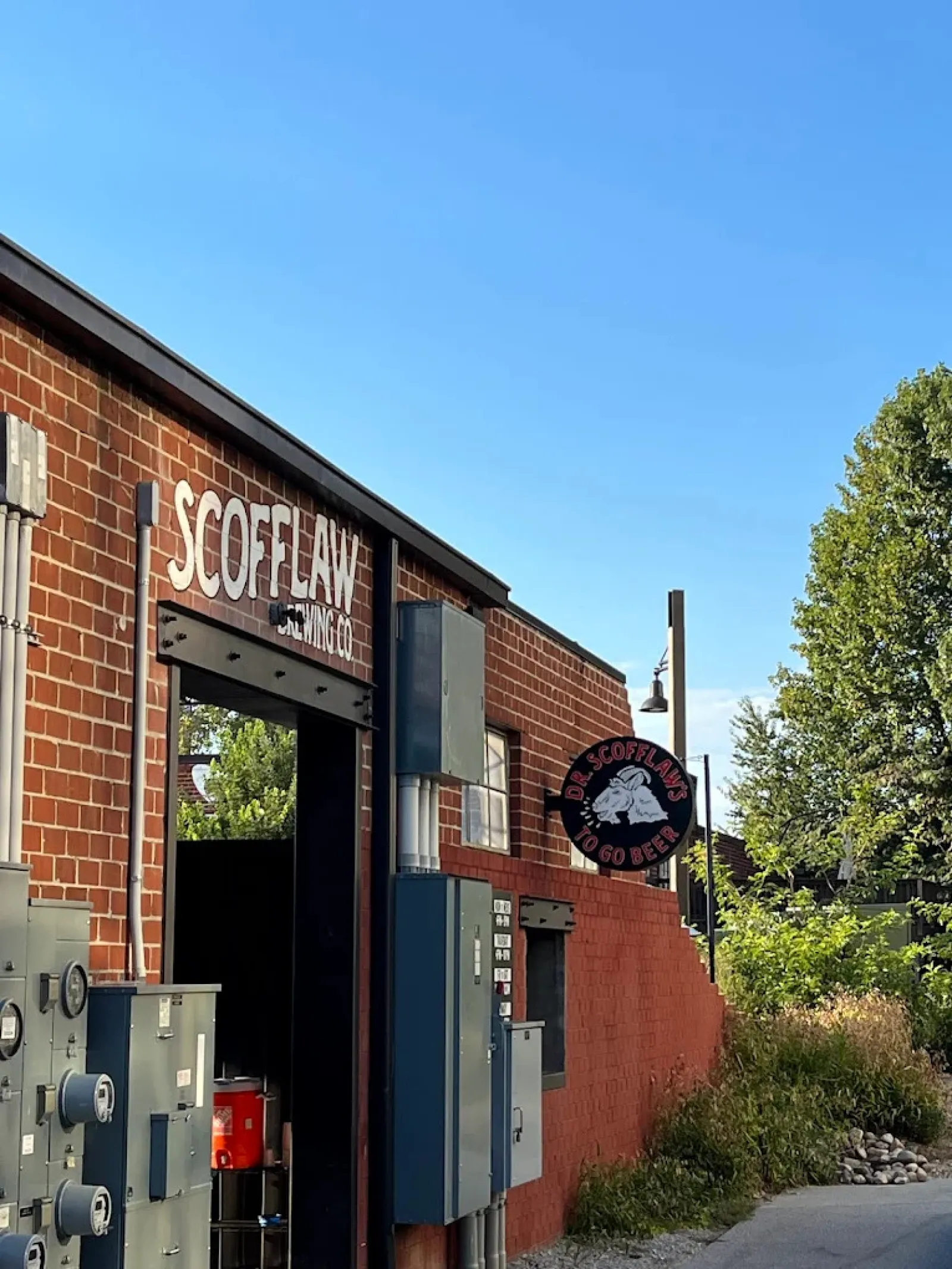 Exterior view of Scofflaw Brewing Co. brick building with signage and lush greenery under clear blue sky.