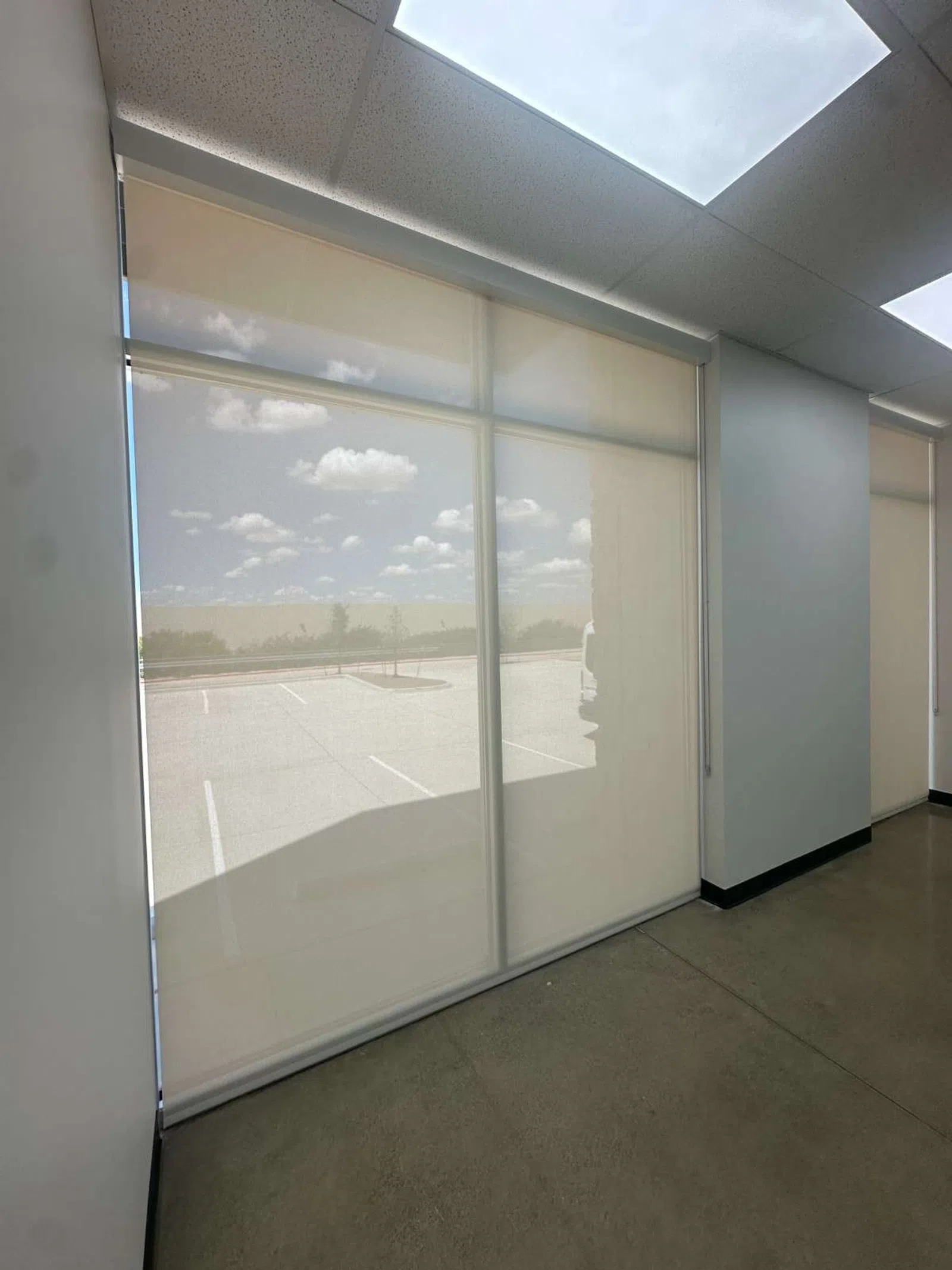 Window with translucent roller shades partially revealing an outdoor parking lot under a cloudy sky.
