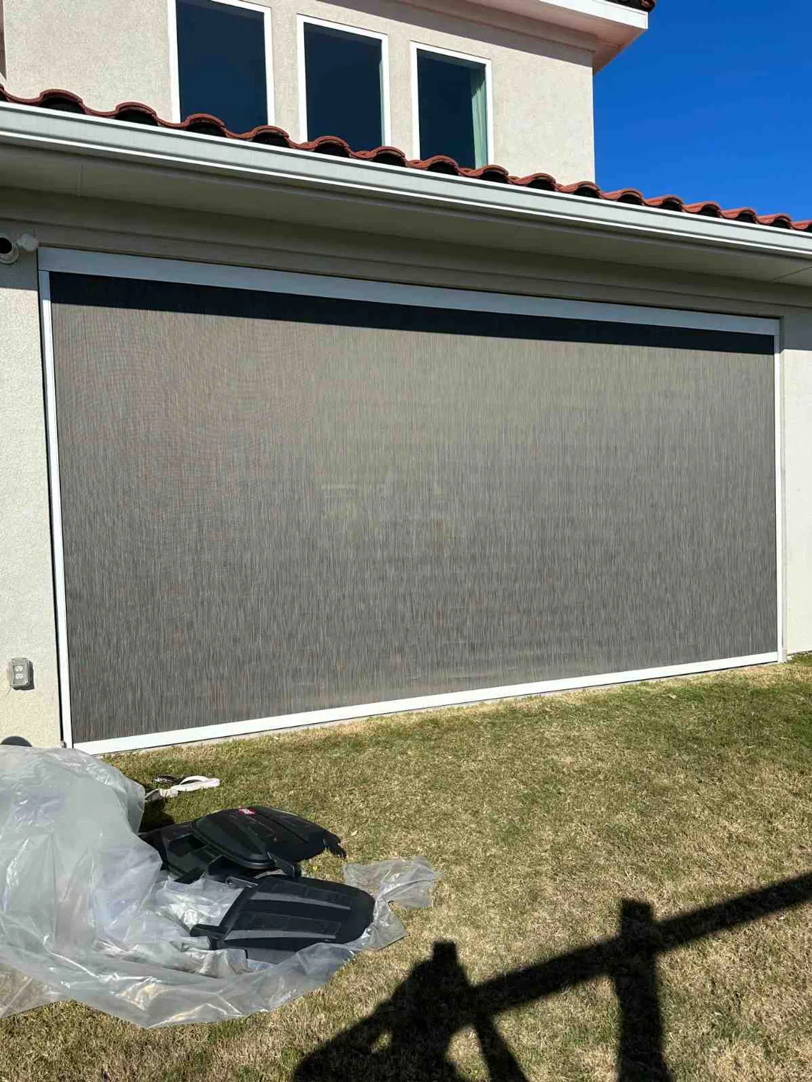 Modern outdoor gray mesh shade screen installed on patio of two-story house with red roof tiles.