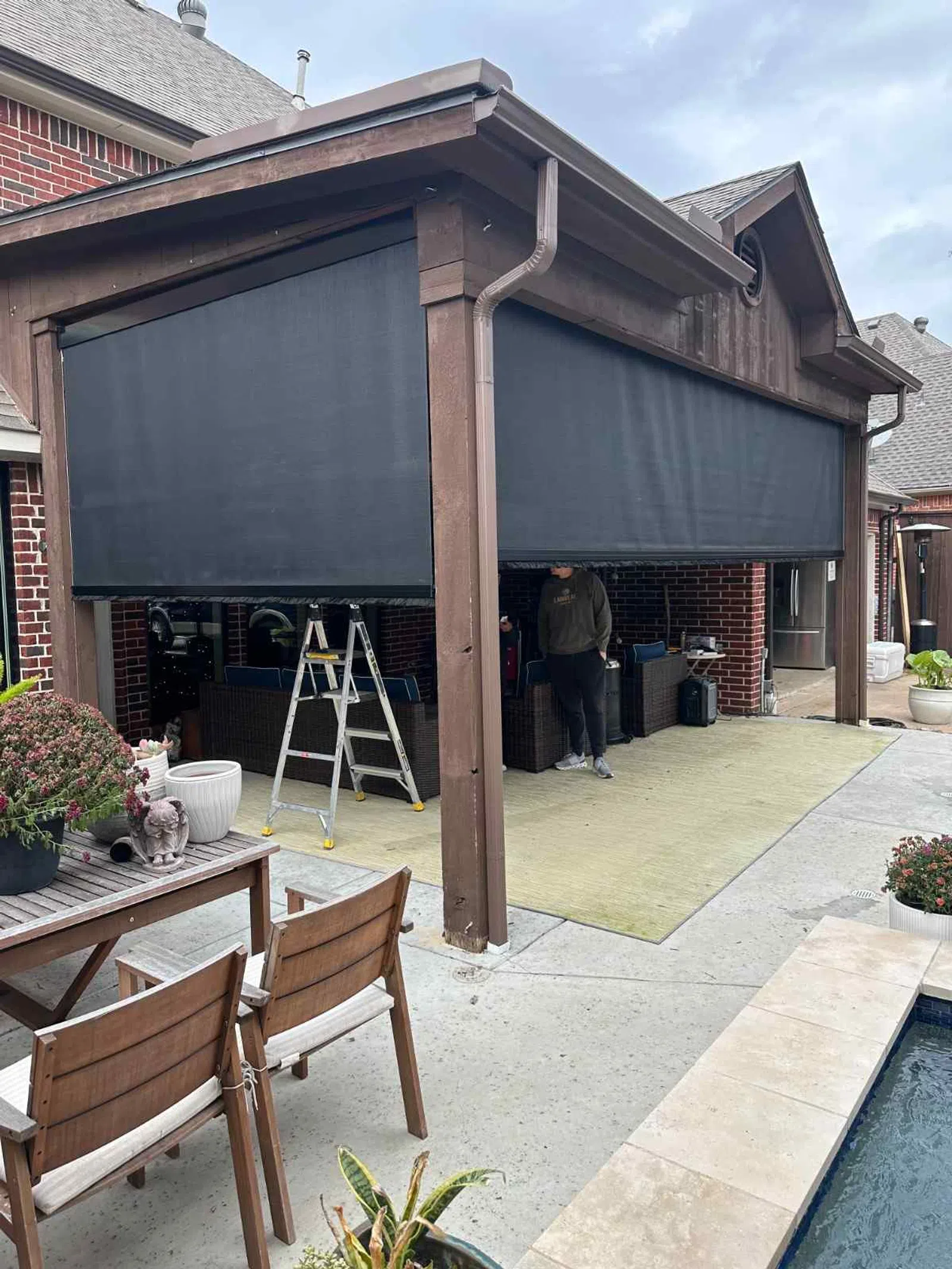 Outdoor covered patio with dark roller shades, wooden furniture, and a swimming pool under cloudy sky.