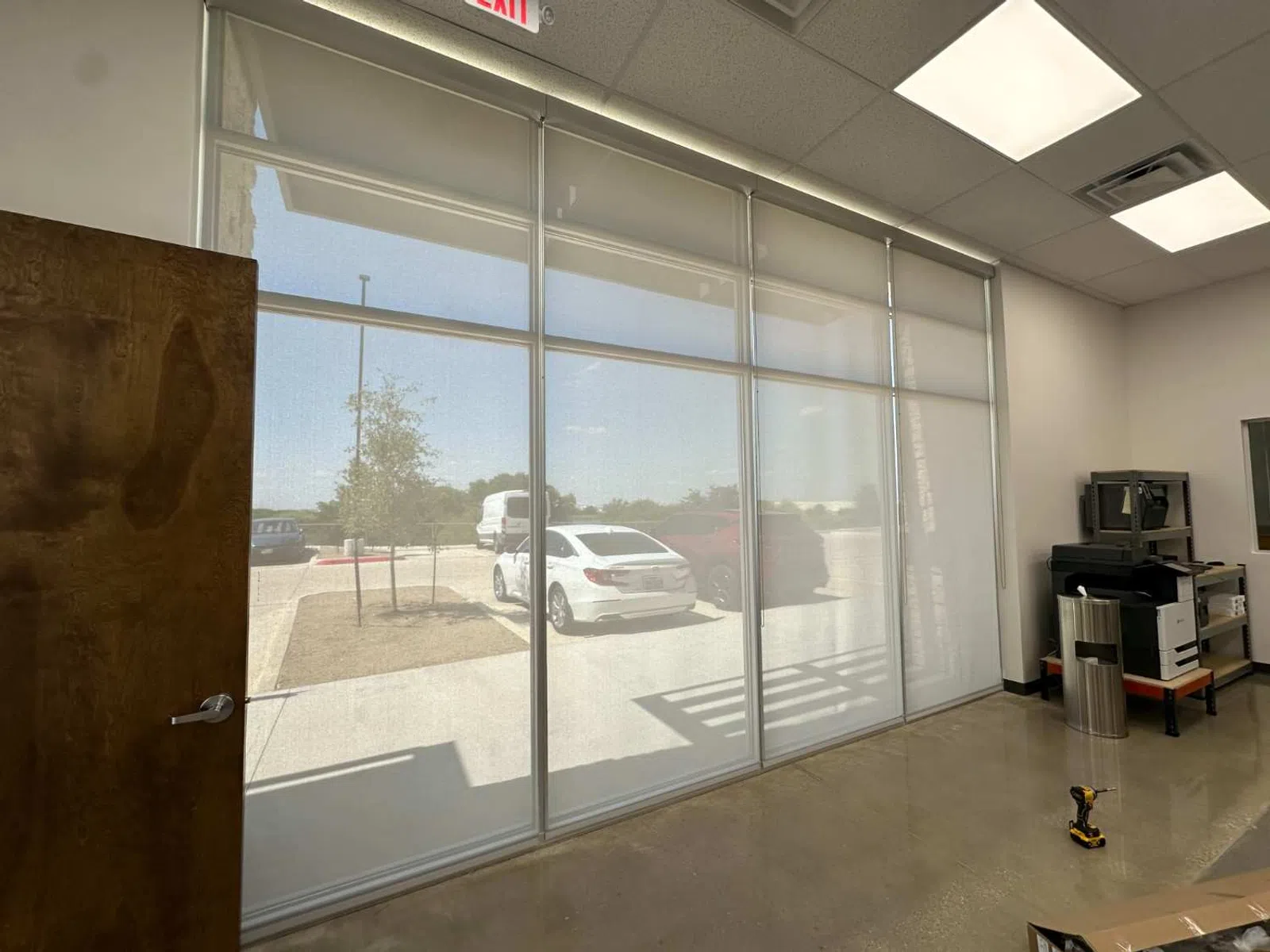 Office interior with large window covered by roller shades showing a parking lot with cars outside.