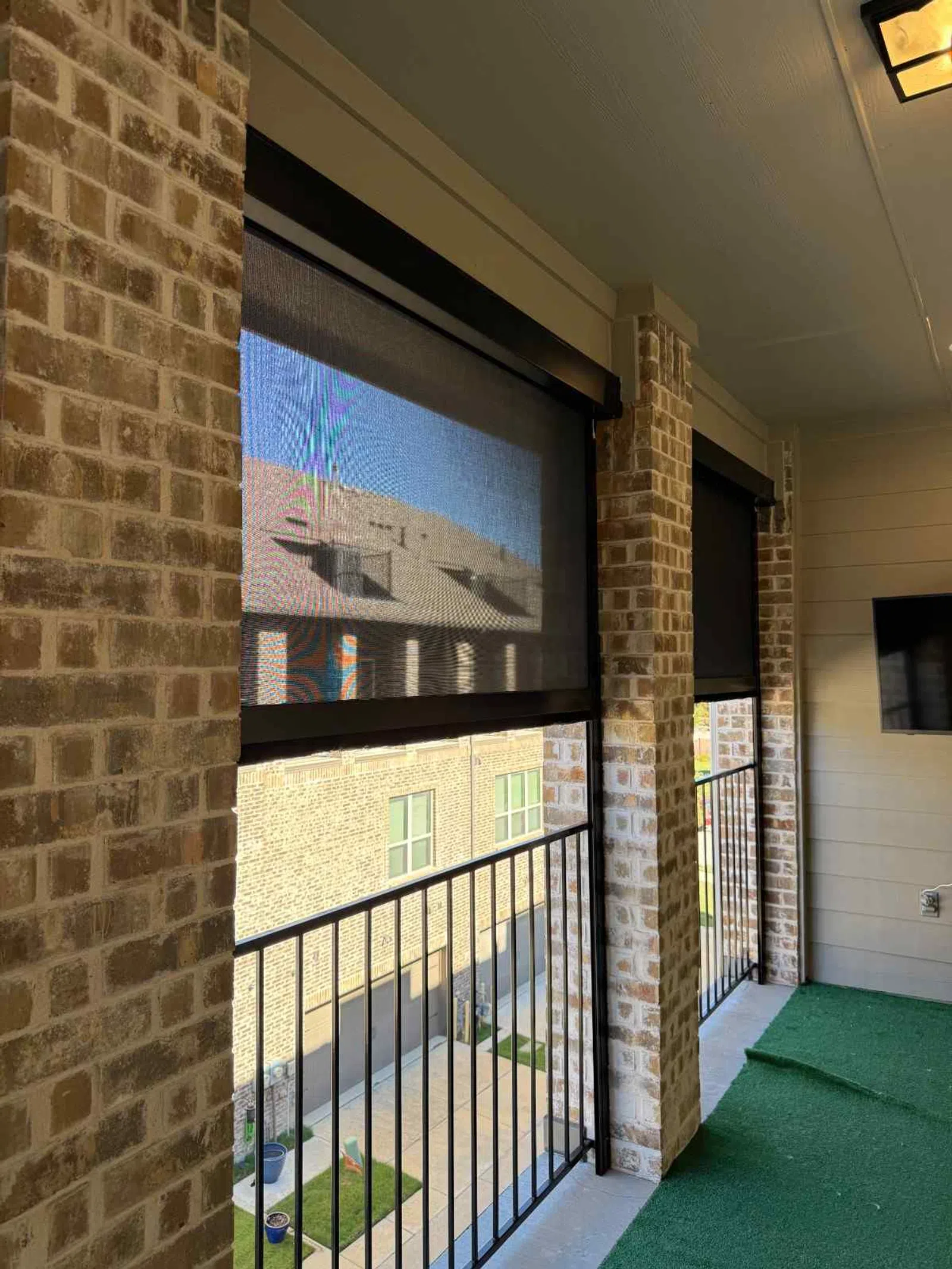 Balcony with brick pillars, black metal railing, green artificial turf floor, and retractable mesh shades under ceiling light.