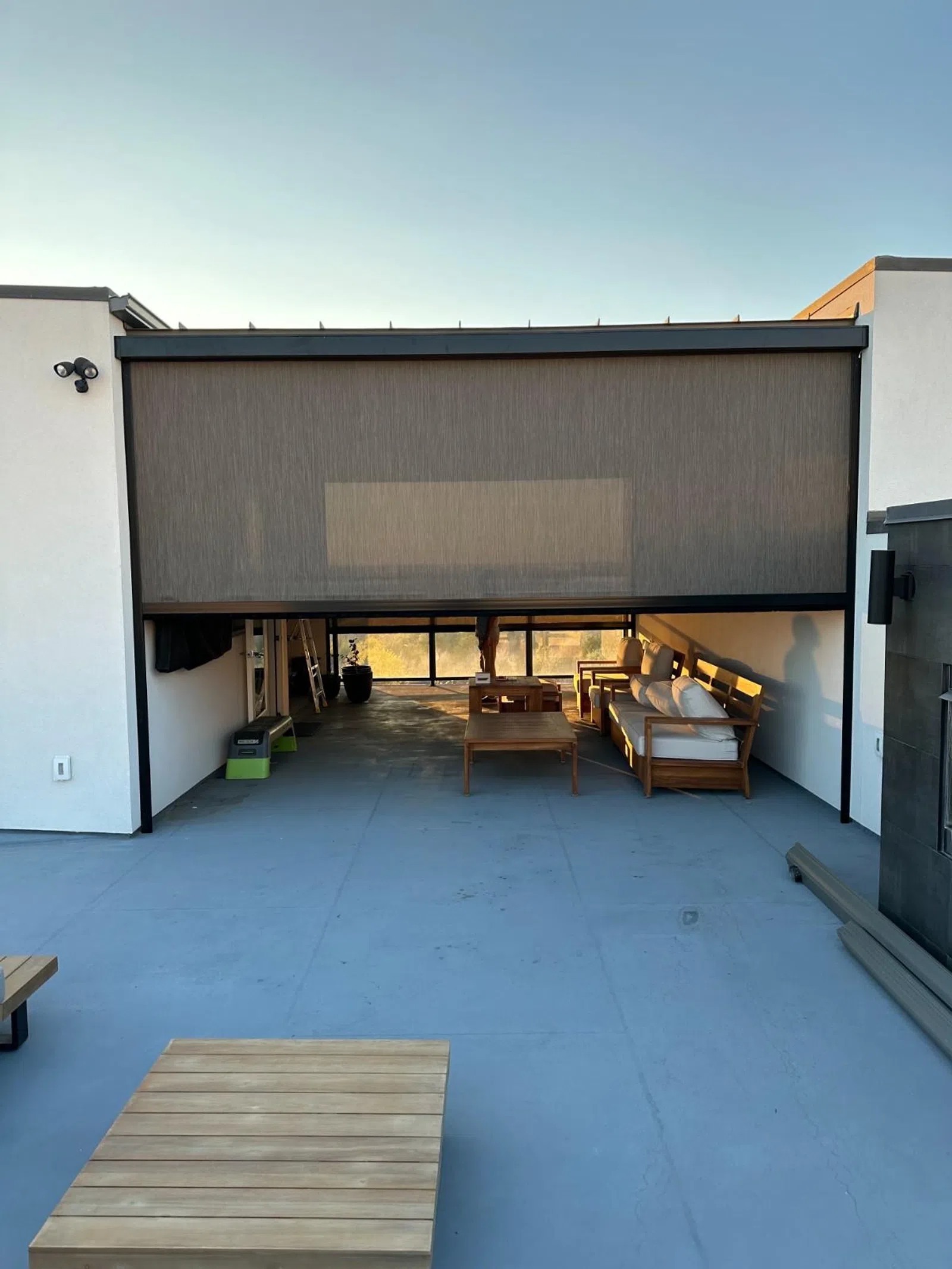 Modern outdoor patio with retractable privacy screen, wooden furniture, and spacious concrete flooring under clear sky.