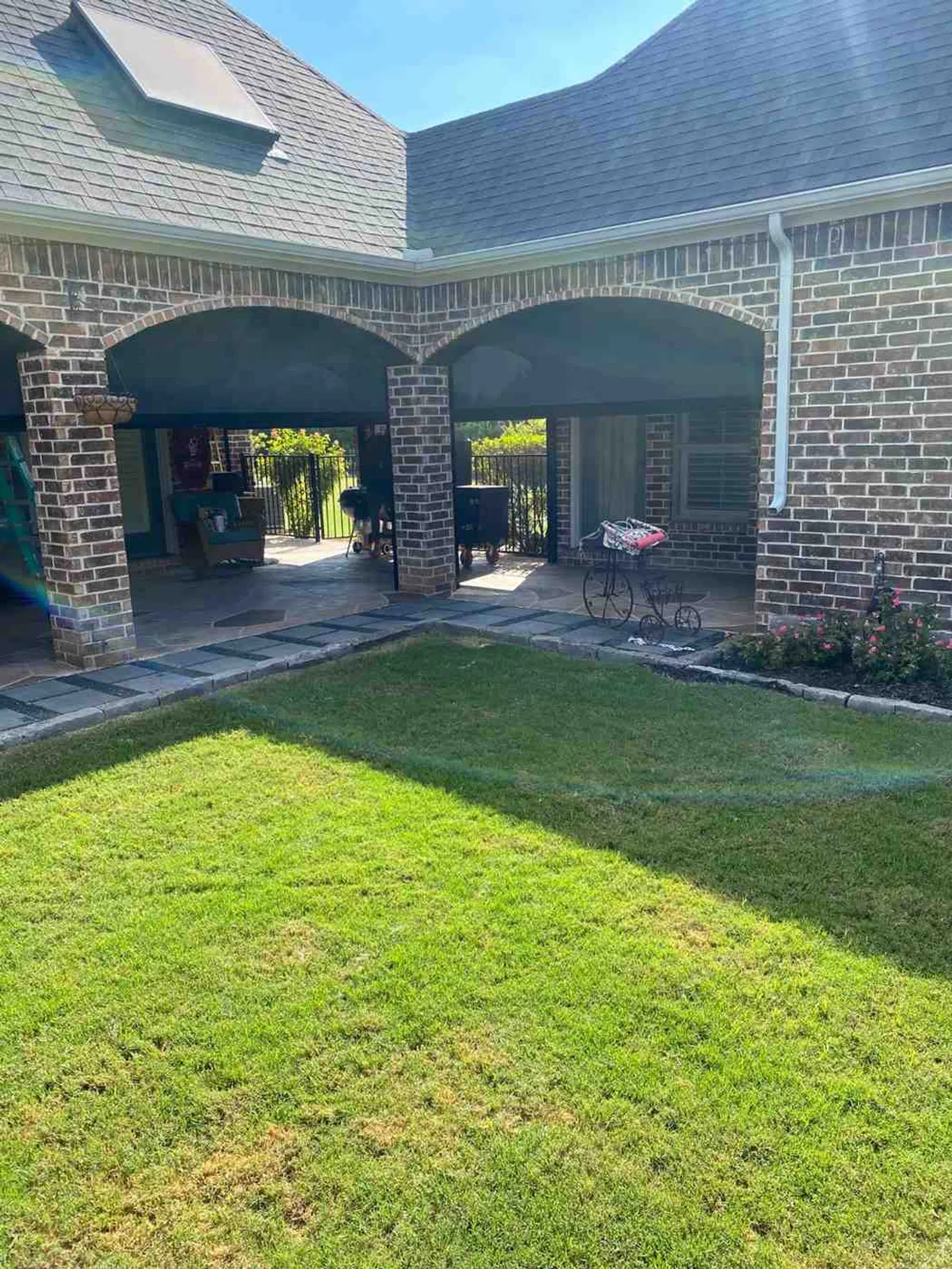Sunny backyard patio with brick arches, neatly trimmed grass, and outdoor furniture in shaded sitting area.