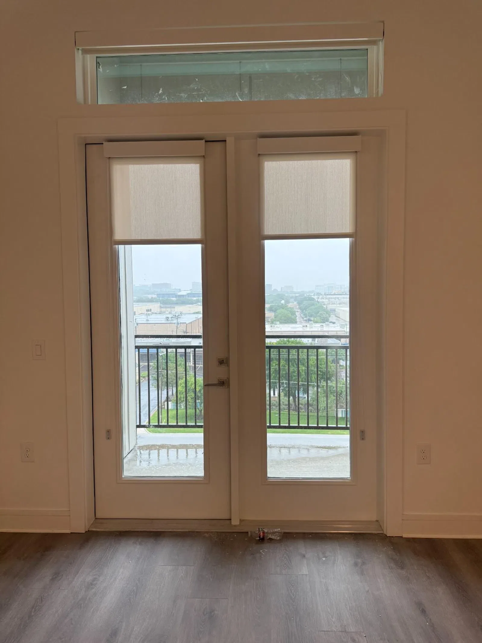 White French doors with partially lowered roller shades open to a balcony overlooking a cityscape on a rainy day.