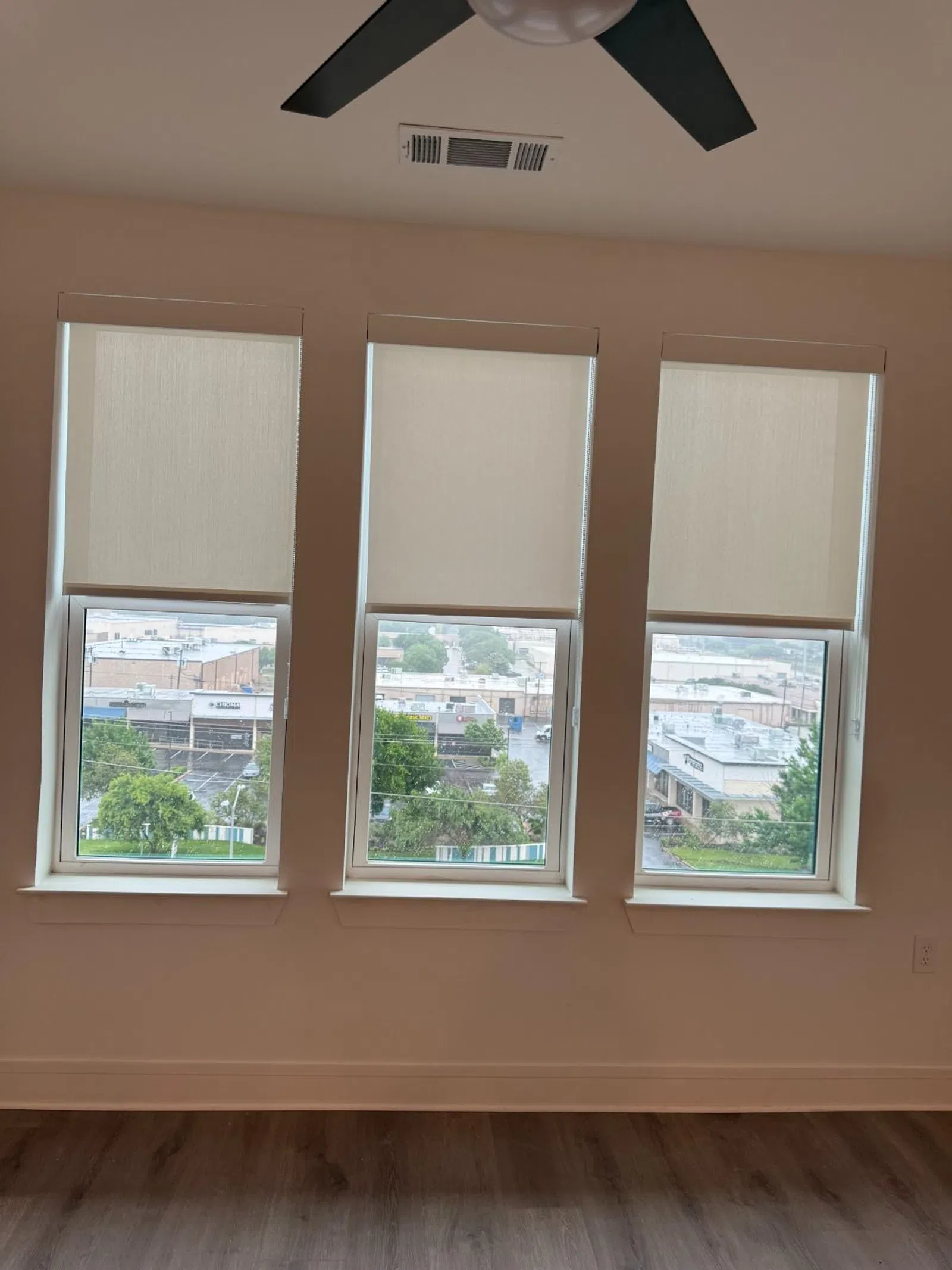 Three windows with beige roller shades partially lowered in a modern room with a ceiling fan and wood flooring.