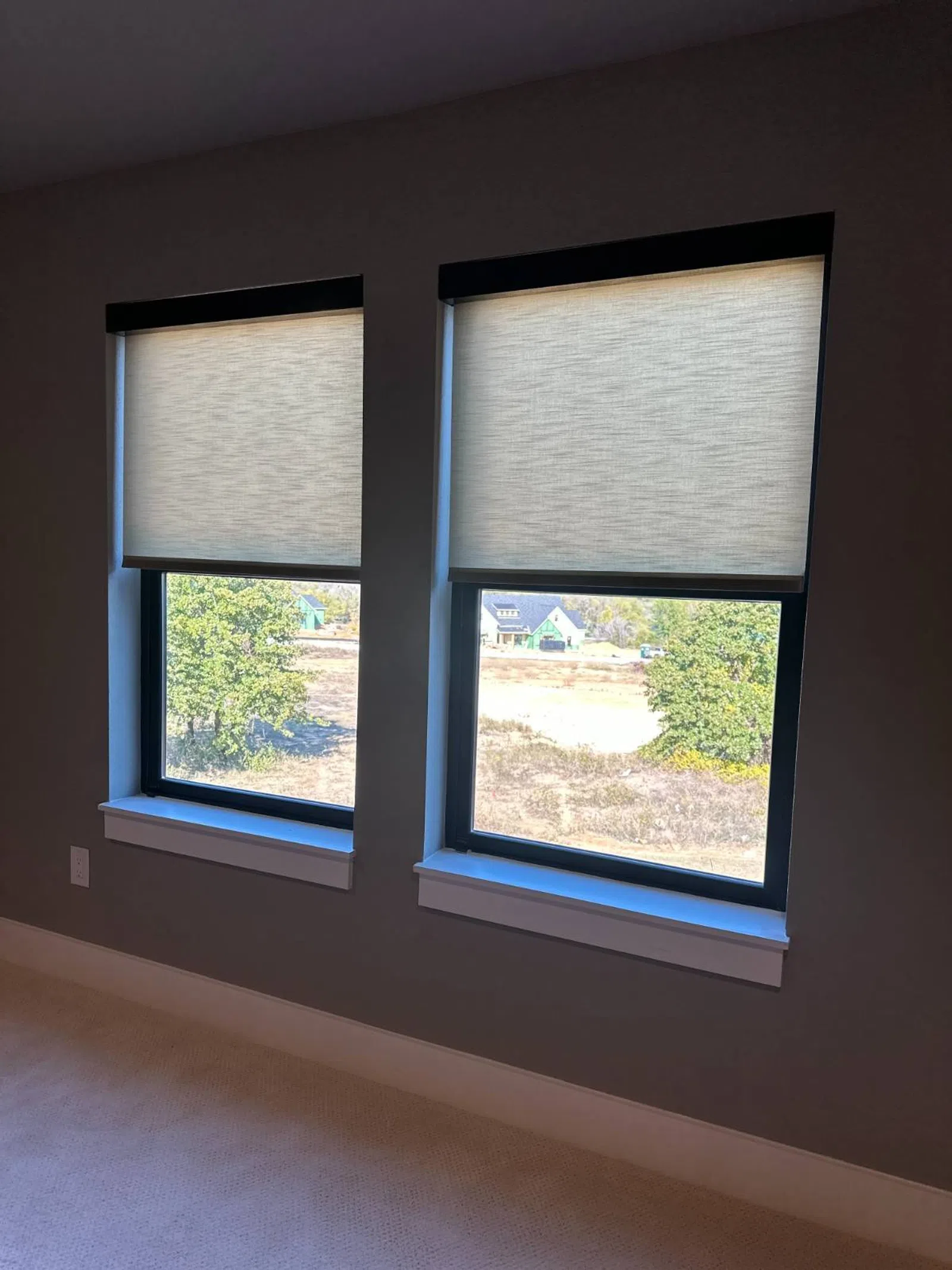 Two windows with beige roller shades partially lowered, showing an outdoor view of trees and houses.