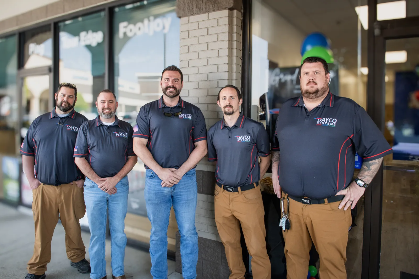 Five men in matching Dayco Systems shirts and khaki pants standing outside a storefront in a row.