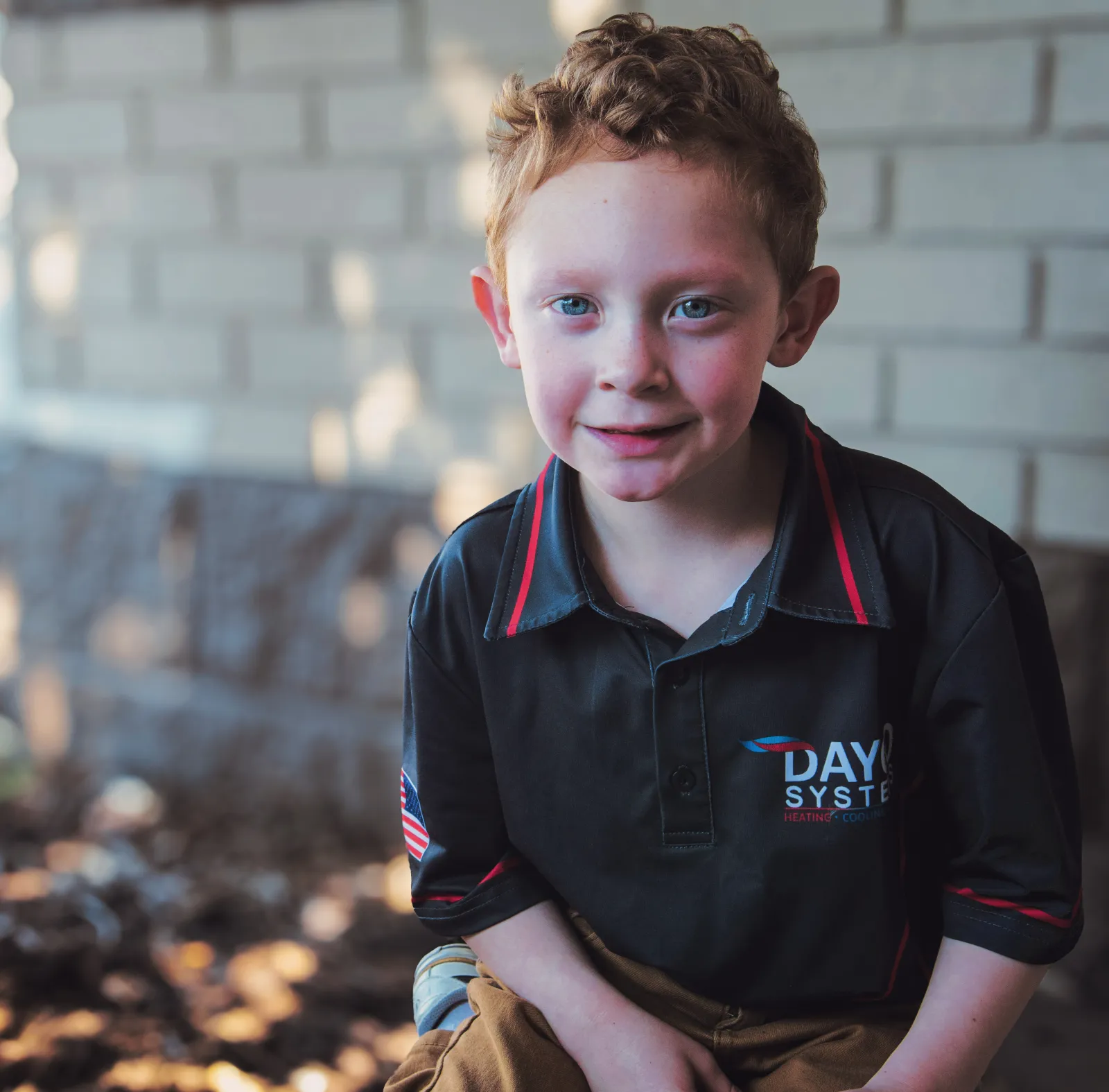 Young boy with curly red hair wearing a black Dayco Systems shirt outdoors near a brick wall.