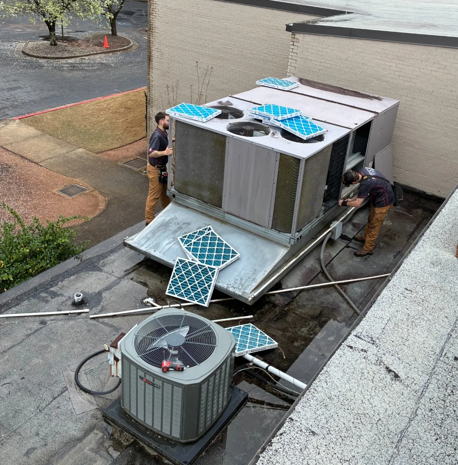 Technicians installing or repairing a rooftop HVAC unit with new blue filters on a flat roof beside a building.
