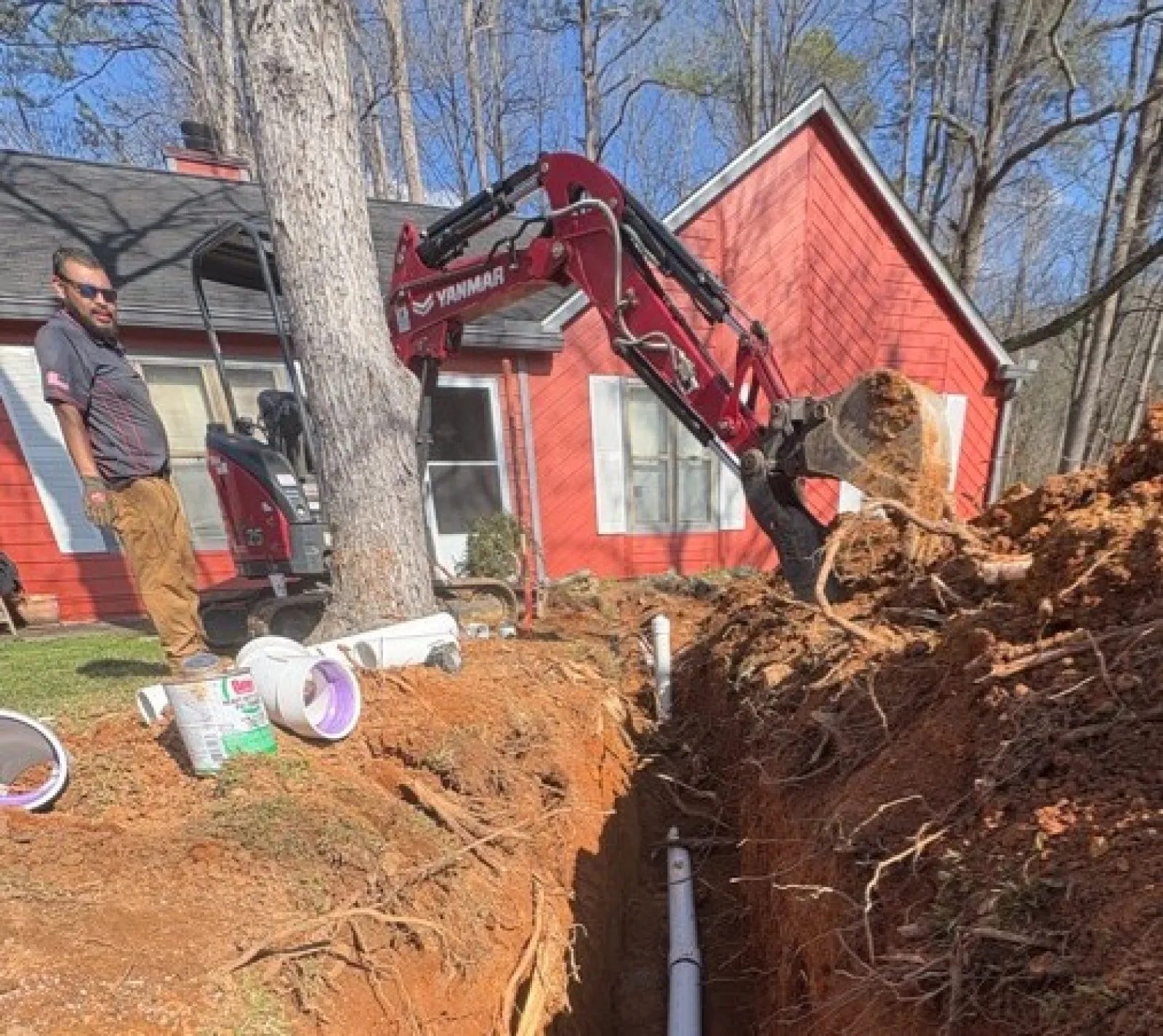 Excavator digging trench for white PVC pipe installation near red house in a wooded area.